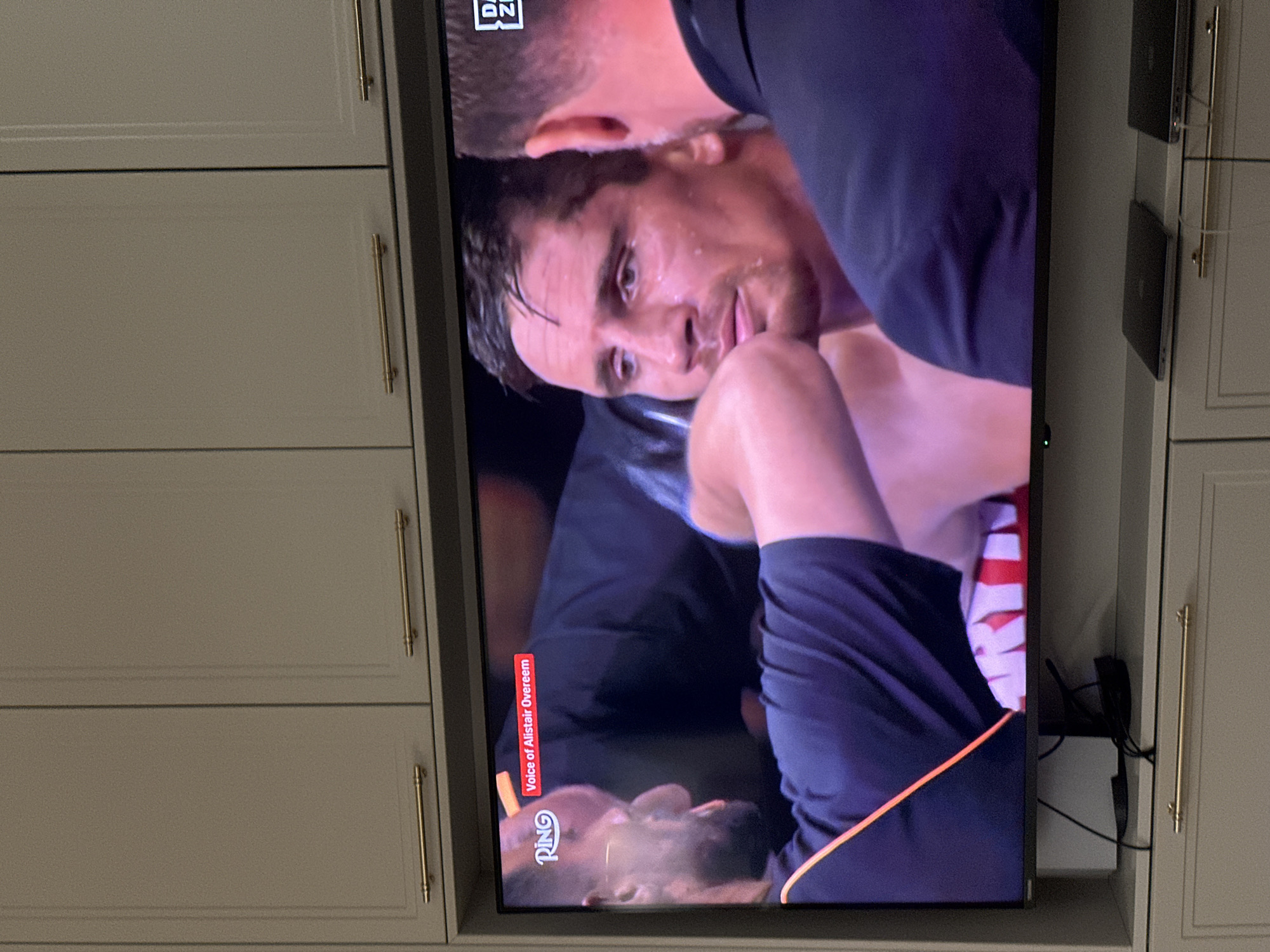 The image shows a television screen displaying a close-up shot of a boxing match. One boxer is leaning over the other who is on the ground looking up. The DAZN logo is visible in the top left corner of the screen. The words "Voice of Alistair Overeem" are visible on the screen. The television is mounted on a wall within a cabinet with long, vertical, gold-colored handles. Two black devices are mounted to the right of the screen, also within the cabinet. The location is unknown.
