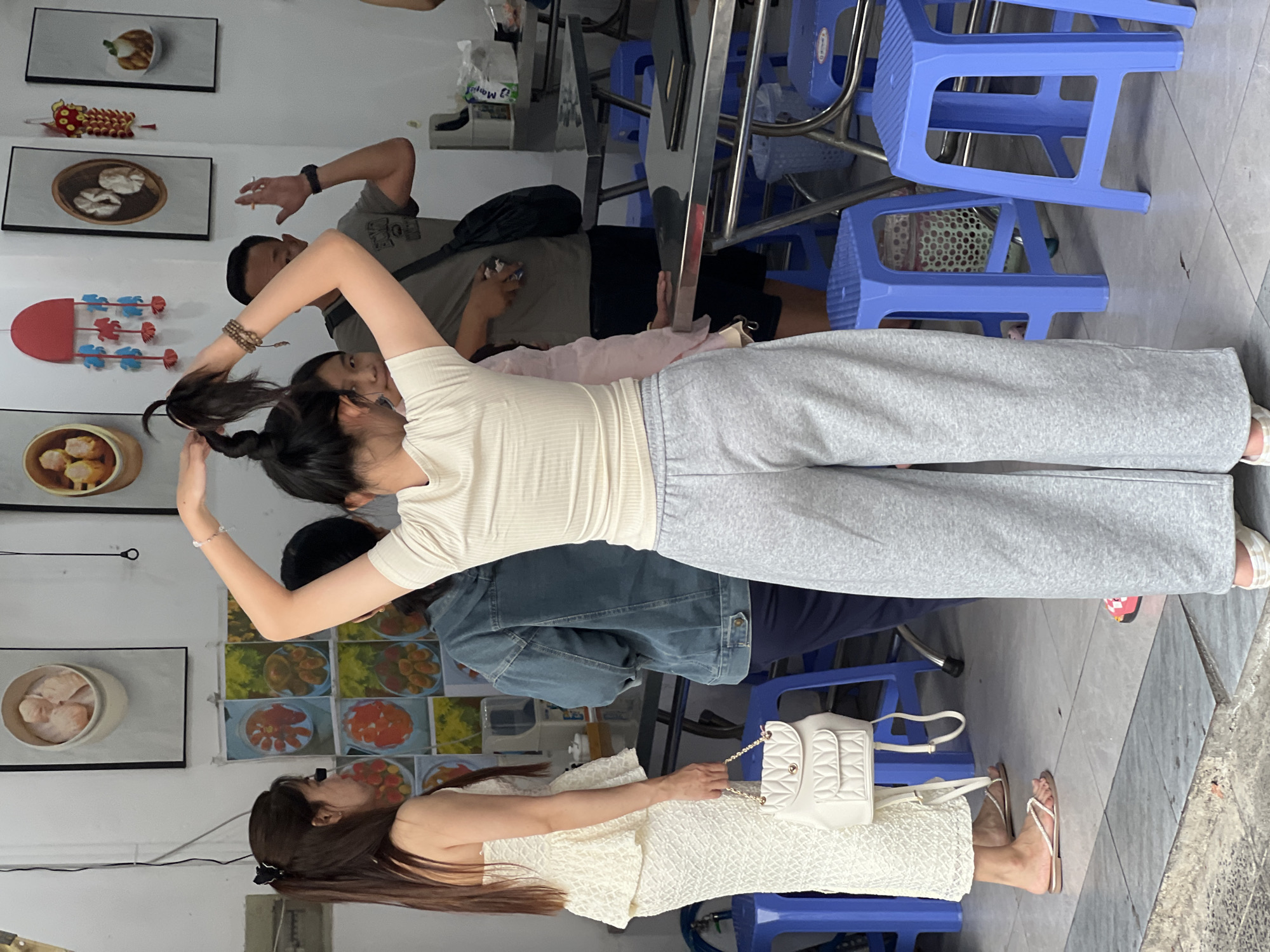 The image depicts an outdoor scene, likely a street-side dining area, in Nha Trang, Vietnam. Several individuals are present. In the foreground, a young woman with long dark hair is styled up, wearing a light-colored ribbed top and grey sweatpants. She is facing away from the camera, with her hands in her hair. To her left, another young woman in a white, textured dress and a white backpack is looking towards the other individuals. Behind them, a man in a grey t-shirt and dark shorts is visible, along with another person wearing a denim jacket. The background features a white wall adorned with framed artwork of food, possibly dim sum. Tables and blue plastic stools are arranged in the space, suggesting a casual eatery. The ground is tiled.