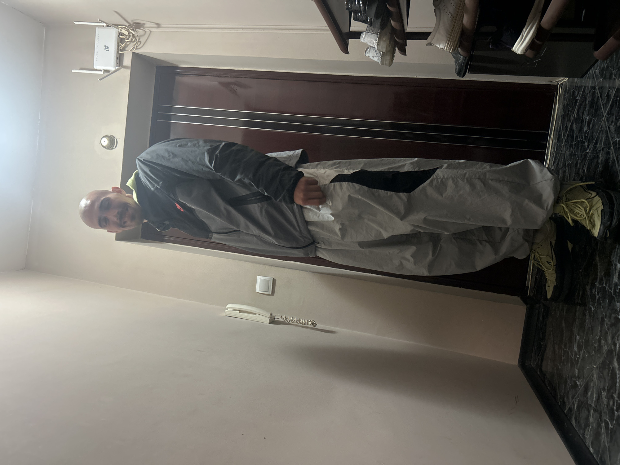A bald man stands in a hallway. He wears a grey jacket and light grey pants with black accents, paired with light green sneakers. To his left, a beige wall is visible, equipped with a landline telephone and a light switch. Above him, a Wi-Fi router is mounted on the wall near a dark wooden door. The hallway floor has a dark, marble-like pattern. The location is possibly Burgas, Bulgaria.
