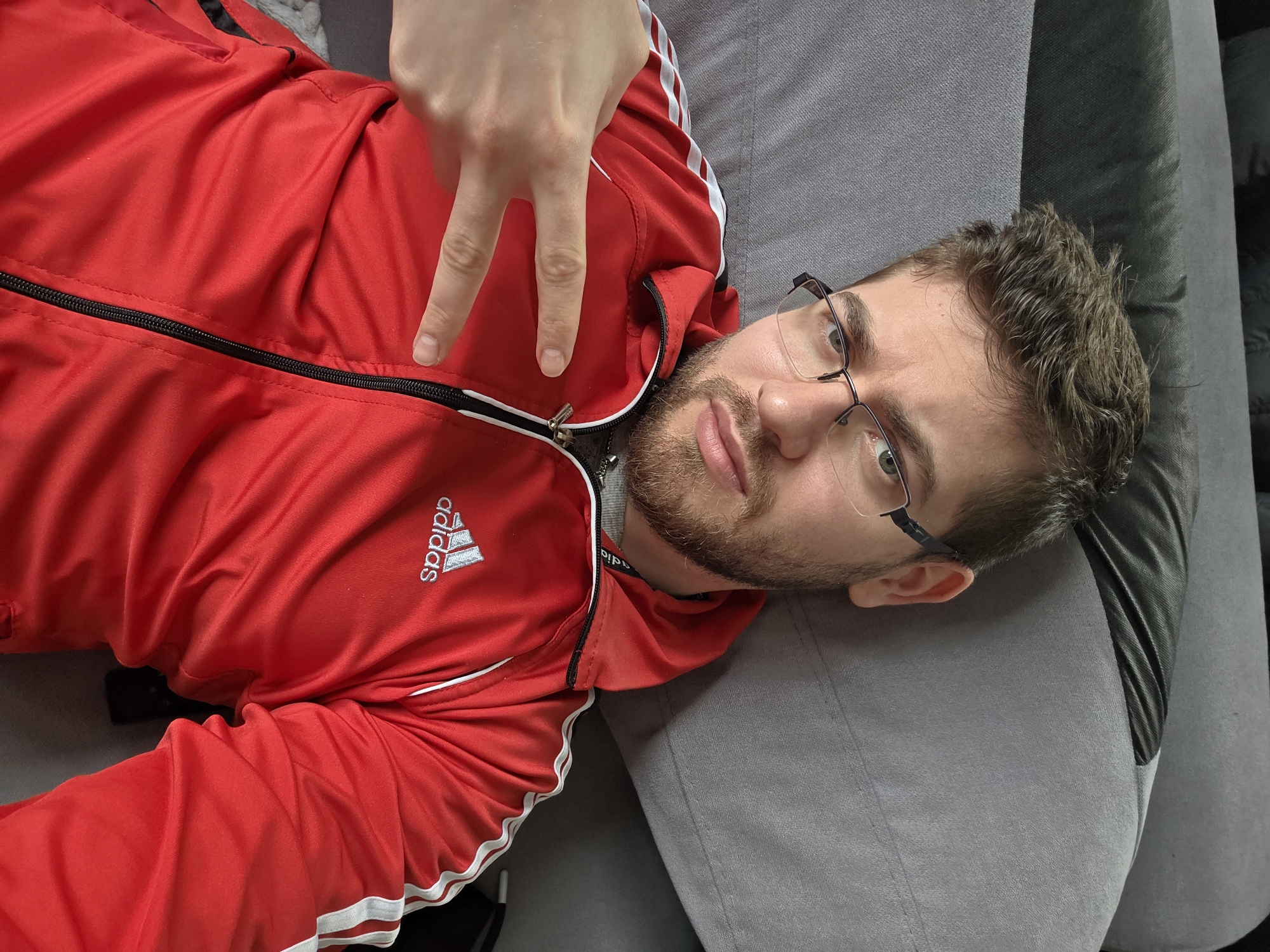 A man is lying on a gray couch. He is wearing a red Adidas jacket with white stripes on the sleeves. He has short brown hair, a beard, and glasses. He is making a peace sign with his left hand. The location is unspecified in Yagodovo, Bulgaria.