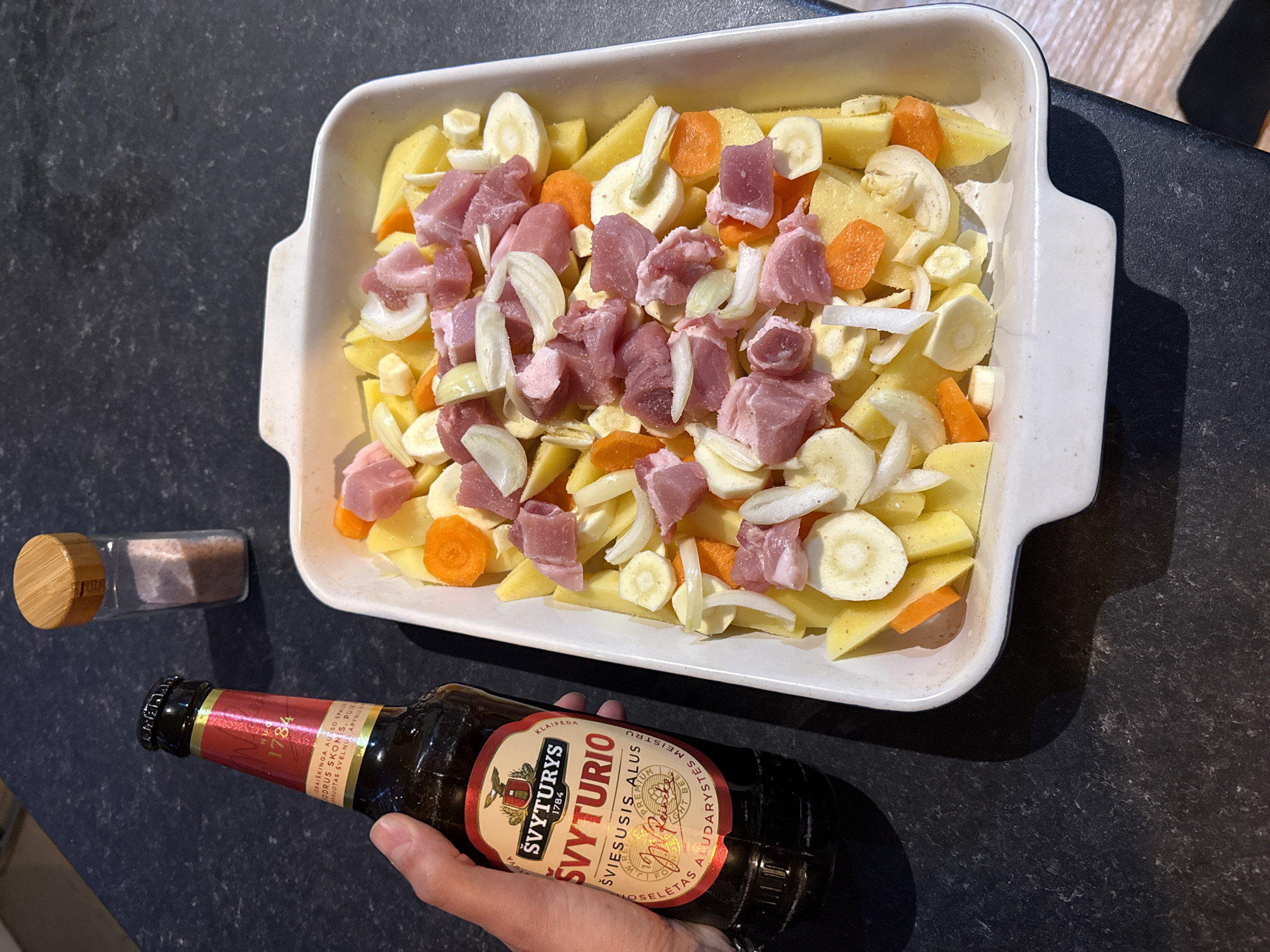 The image depicts a culinary scene, likely taking place indoors on a dark countertop. A white, rectangular baking dish filled with chopped vegetables, including potatoes, carrots, onions, and parsnips, is positioned at the top. On top of the vegetables, there are pieces of raw pork. To the left of the baking dish is a small glass container with a wooden lid, possibly holding salt or spices.
In the foreground, a hand is visible holding a brown glass bottle of Švyturys beer. The label is clearly visible, showing the brand name and other details. The overall composition suggests a meal preparation or cooking process. The image does not provide any details connecting the scene to Great Yarmouth.