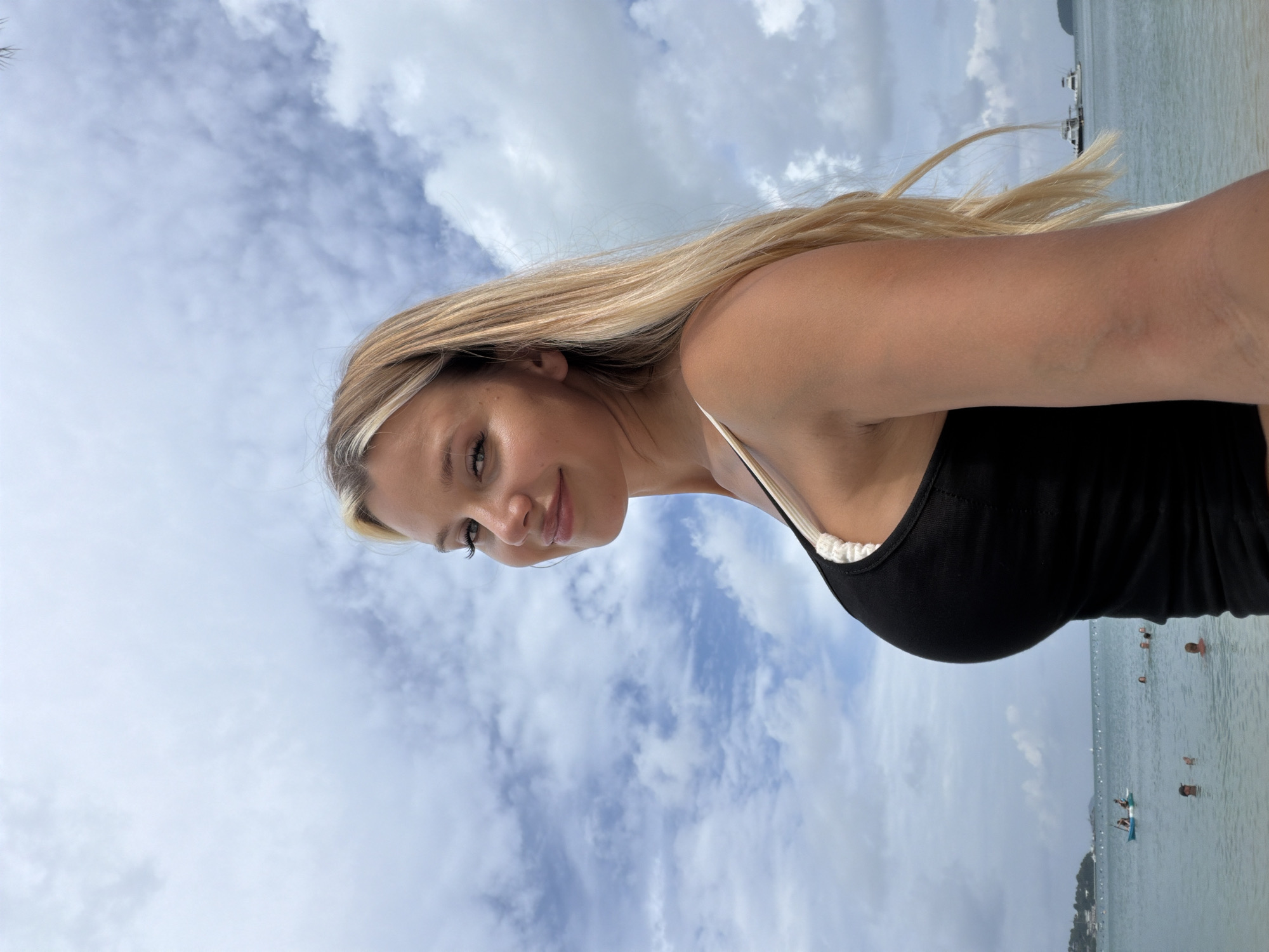 A blonde woman in a black tank top stands on a beach in Wichit, Thailand, looking over her shoulder at the camera. She has long, blonde hair and is smiling. The ocean is in the background, with several boats and people swimming. The sky is cloudy.