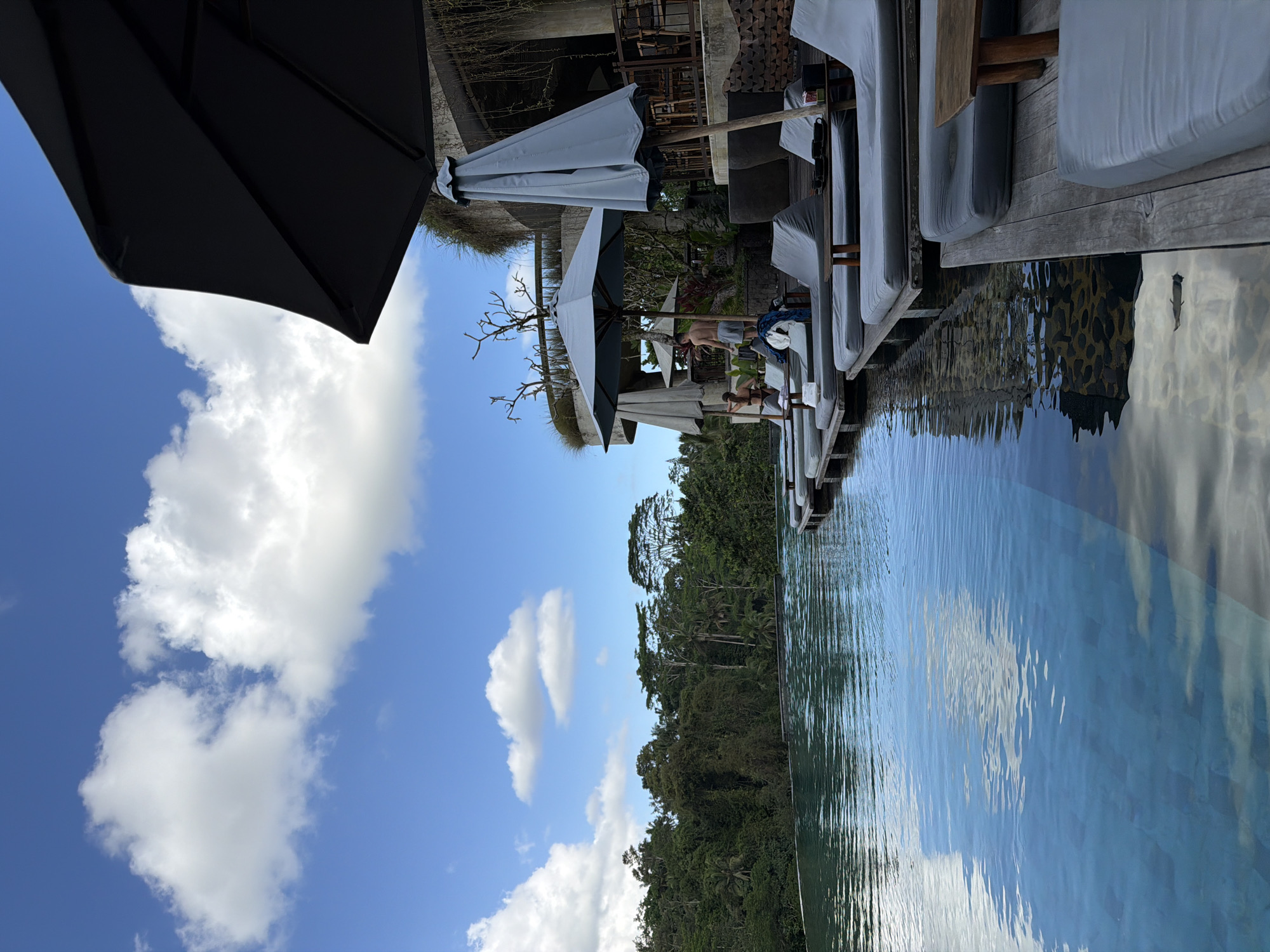 This is a daytime, eye-level, medium shot photograph of an infinity pool overlooking a dense, green jungle in Tegallalang, Indonesia. The sky above is a vibrant blue, dotted with fluffy white clouds. On the right side of the frame, a large dark gray umbrella is angled downwards, partially obscuring the view of a multi-story building constructed with natural materials like stone and wood. Below the umbrella, a smaller, light gray umbrella stands open.
In front of the building, a series of gray lounge chairs are arranged on a wooden deck, with some occupied by people. One man, wearing gray shorts, stands near the edge of the pool, while a woman in a bikini sits on a lounge chair. Another lounge chair in the foreground is empty. The infinity pool itself occupies the left side of the image, its blue water reflecting the sky and clouds, with a tiled bottom visible in the shallower areas. Beyond the pool, the lush, green jungle stretches to the horizon, creating a dramatic backdrop. The scene conveys a sense of relaxation and natural beauty.