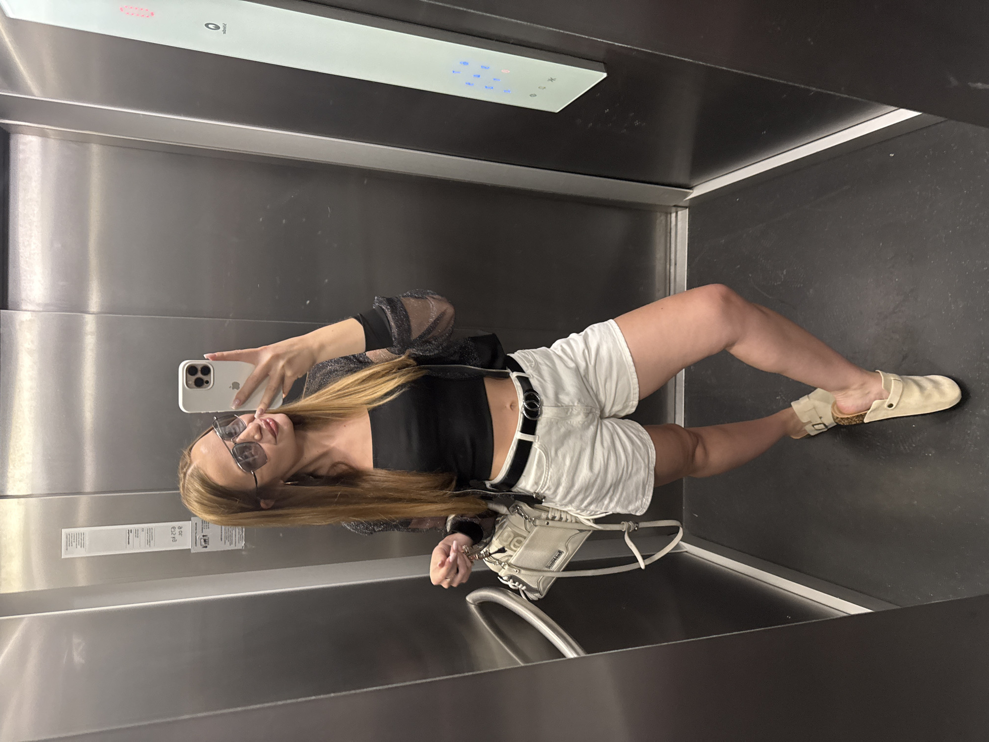 A young woman in a black top, sheer jacket, white shorts, and beige clogs takes a selfie in an elevator. She holds a white phone in her right hand and a white purse in her left. She wears sunglasses. The elevator has stainless steel walls and a black floor. Elevator buttons are visible above her. The location is an elevator in Budva, Montenegro.