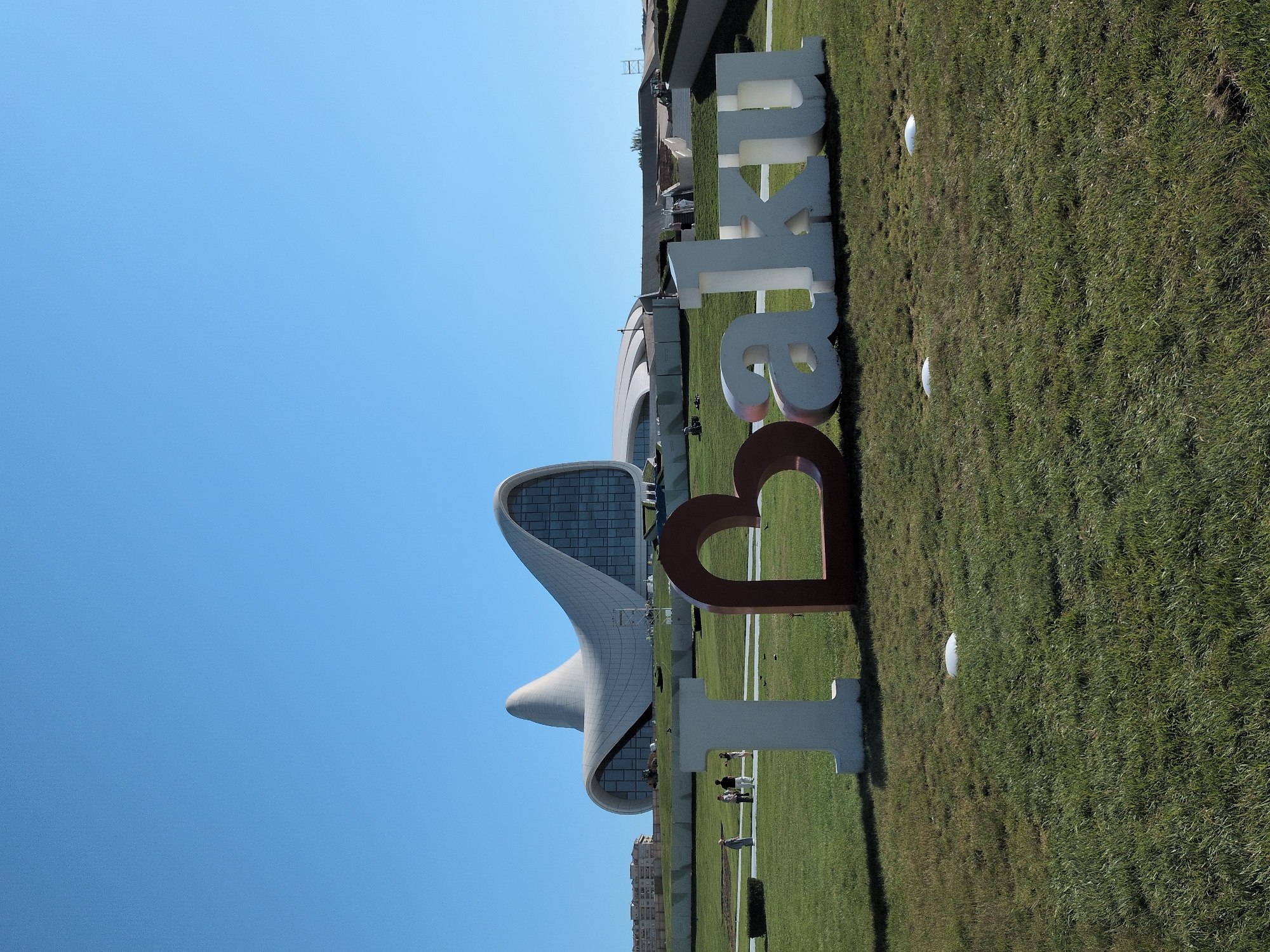 The image depicts a clear, sunny day with a bright blue sky over a grassy lawn in Baku, Azerbaijan. Prominently displayed in the foreground are large, three-dimensional letters spelling out "I Love Baku." The "I" is white, the "Love" is represented by a large, reddish-brown heart shape, and "Baku" is spelled out in white letters.
In the background, the iconic Heydar Aliyev Center, a distinctive white, curved architectural masterpiece, dominates the skyline. To the left of the Heydar Aliyev Center, several smaller buildings are visible. A few people are scattered across the lawn, some walking and others sitting. The environment appears to be a well-maintained park or public space, with manicured grass and some landscaping features.