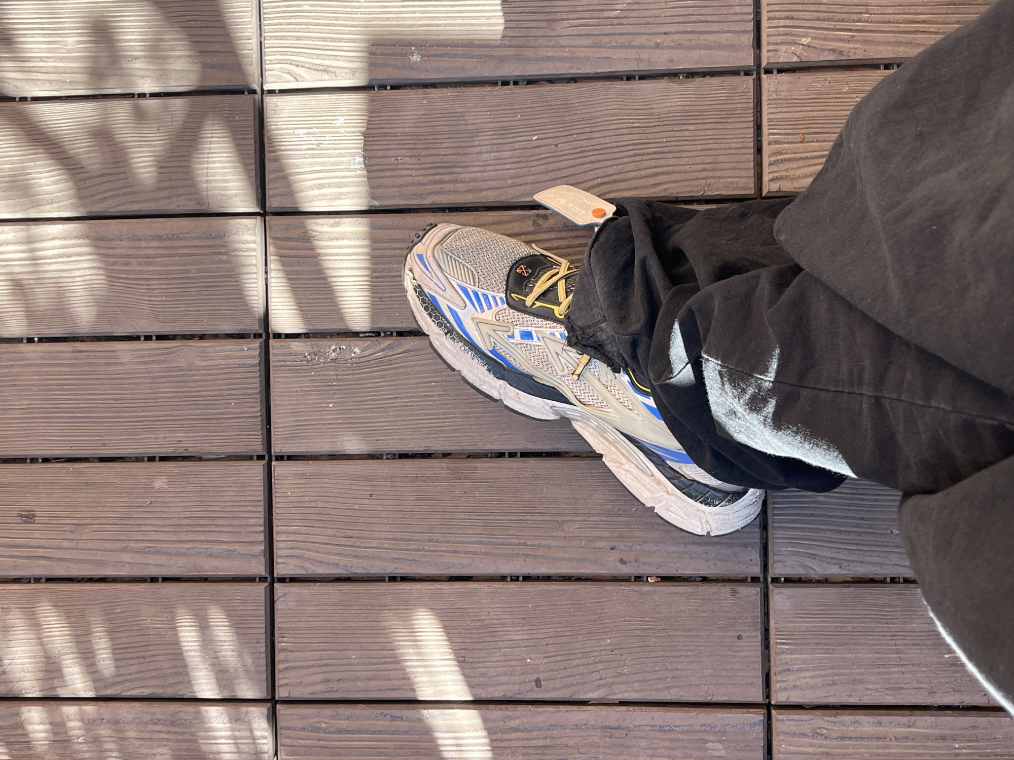 The image shows a person's foot wearing a grey and white athletic shoe with yellow laces and a blue accent. The shoe has a tag attached. The person is also wearing black sweatpants. The person is standing on a wooden deck with parallel planks. The planks appear to be aged and have parallel wood grain. The lighting on the deck is uneven, with strong shadows cast across the planks. There is no information from the image that indicates the image was taken in Barcelona, Spain.