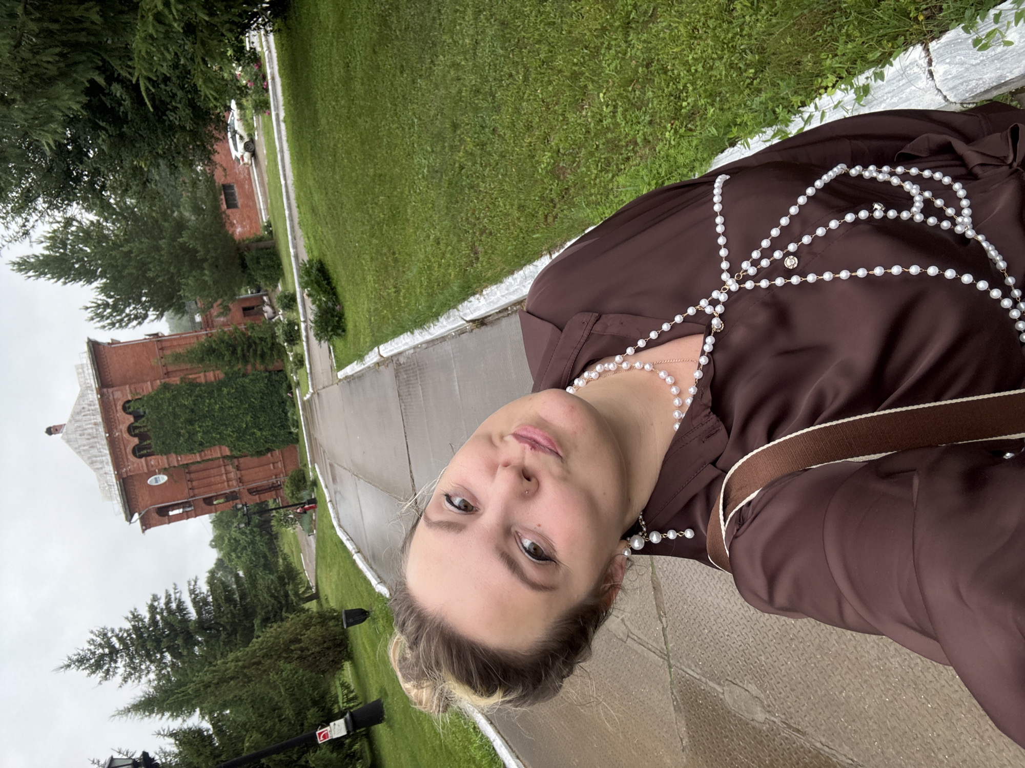 This is a vertical, slightly tilted, eye-level selfie of a young woman taken outdoors. The woman, who appears to be Caucasian, has blonde hair pulled back into a bun and is wearing a brown, sheer, long-sleeved dress adorned with a decorative pearl necklace. She has on pearl earrings as well. She is looking directly at the camera with a neutral expression. A brown strap, possibly from a bag, crosses her shoulder.
The background shows a paved walkway, a well-maintained grassy area, and in the distance, a red brick building that appears to be an old church or historical structure, partially covered in ivy. Trees and other greenery are also visible. The lighting suggests it might be daytime, possibly overcast due to the muted light. The wet appearance of the walkway could indicate recent rain. The location is identified as Zavod Mikhailovskii, Russia. There is no visible text in the image.