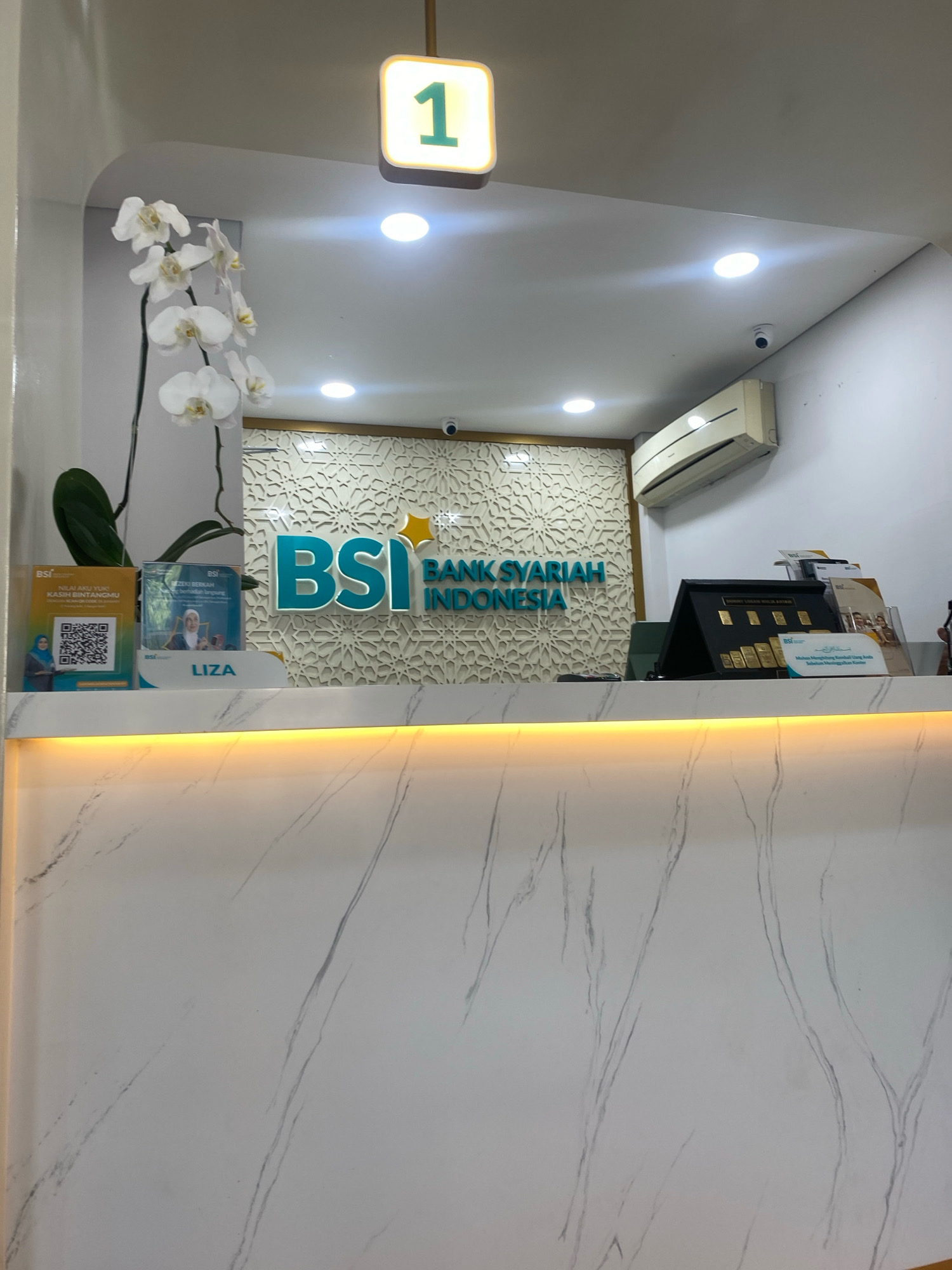 The image depicts the interior of a Bank Syariah Indonesia (BSI) branch, likely located in Banjarbaru, Indonesia. The focal point is a counter area with a large, illuminated BSI logo behind it. Above the counter, a digital display shows the number "1" in green.
On the left side of the counter, there are two brochures or pamphlets displayed. One appears to be a QR code for "NILAI AKU YUK KASIH BINTANGMU", and the other features a woman's portrait and the text "LIZA". A white orchid plant with several blooms is positioned to the left of these brochures.
The counter itself is a clean, white surface with a subtle gray marble-like pattern. A warm yellow light emanates from underneath the counter, illuminating the front of the counter.
To the right of the BSI logo, there's a display stand holding several gold bars, suggesting the bank offers gold investment services. A black stand is used to showcase these bars. Additional brochures are placed to the right of the gold bar display.
In the background, the wall behind the counter features a decorative, light-colored geometric pattern. An air conditioning unit is visible mounted high on the right wall, and a small security camera is also present on the ceiling. The overall impression is one of a modern, well-lit, and professional banking environment.