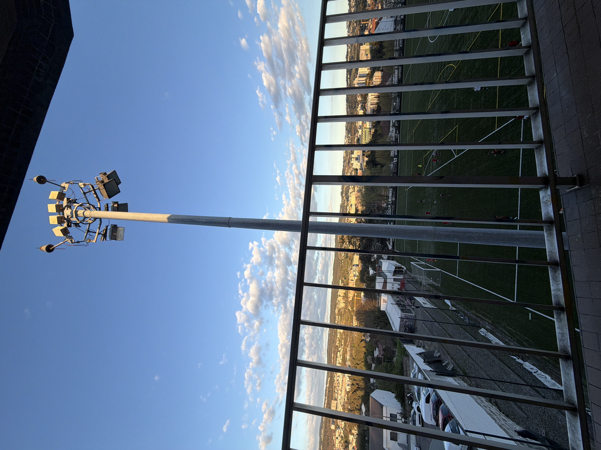 The image depicts a view from a high vantage point, possibly a balcony, in Vila Real, Portugal. A metal railing runs horizontally across the frame. Beyond the railing, the scene is divided into two distinct areas. To the left, a tall light pole with multiple spotlights extends upward into a blue sky with scattered clouds. To the right, a soccer field is visible, with players engaged in what appears to be a practice or game. Buildings and trees are visible in the background. The lighting suggests it is either early morning or late afternoon. Several vehicles are parked beneath.