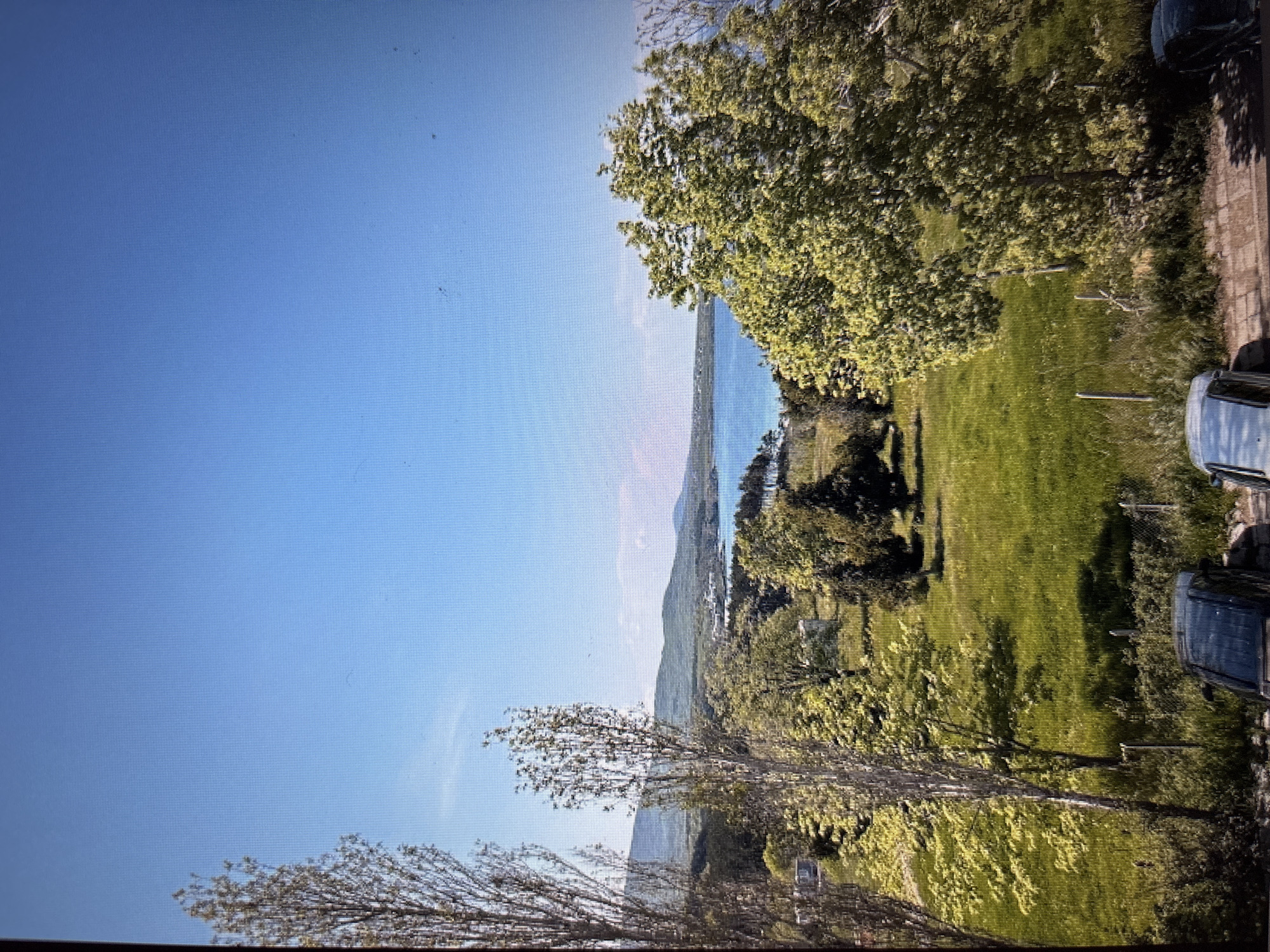 The image depicts a bright, clear day with a vast expanse of blue sky. Below the sky, a serene body of blue water, likely a bay or sea, is visible, bordered by a green, hilly coastline. In the foreground, a vibrant green meadow is dotted with trees of varying sizes and foliage density. To the left, tall, slender trees with sparse leaves frame the scene. The meadow also contains some man-made structures, possibly small buildings or sheds. In the lower part of the image, a paved area shows two dark-colored cars parked side-by-side. A wire fence with wooden posts runs across the foreground. The overall impression is a peaceful, natural landscape with hints of human presence. This scene is not located in Sofia, Bulgaria, as the environment suggests a coastal or lakeside setting rather than an urban one.