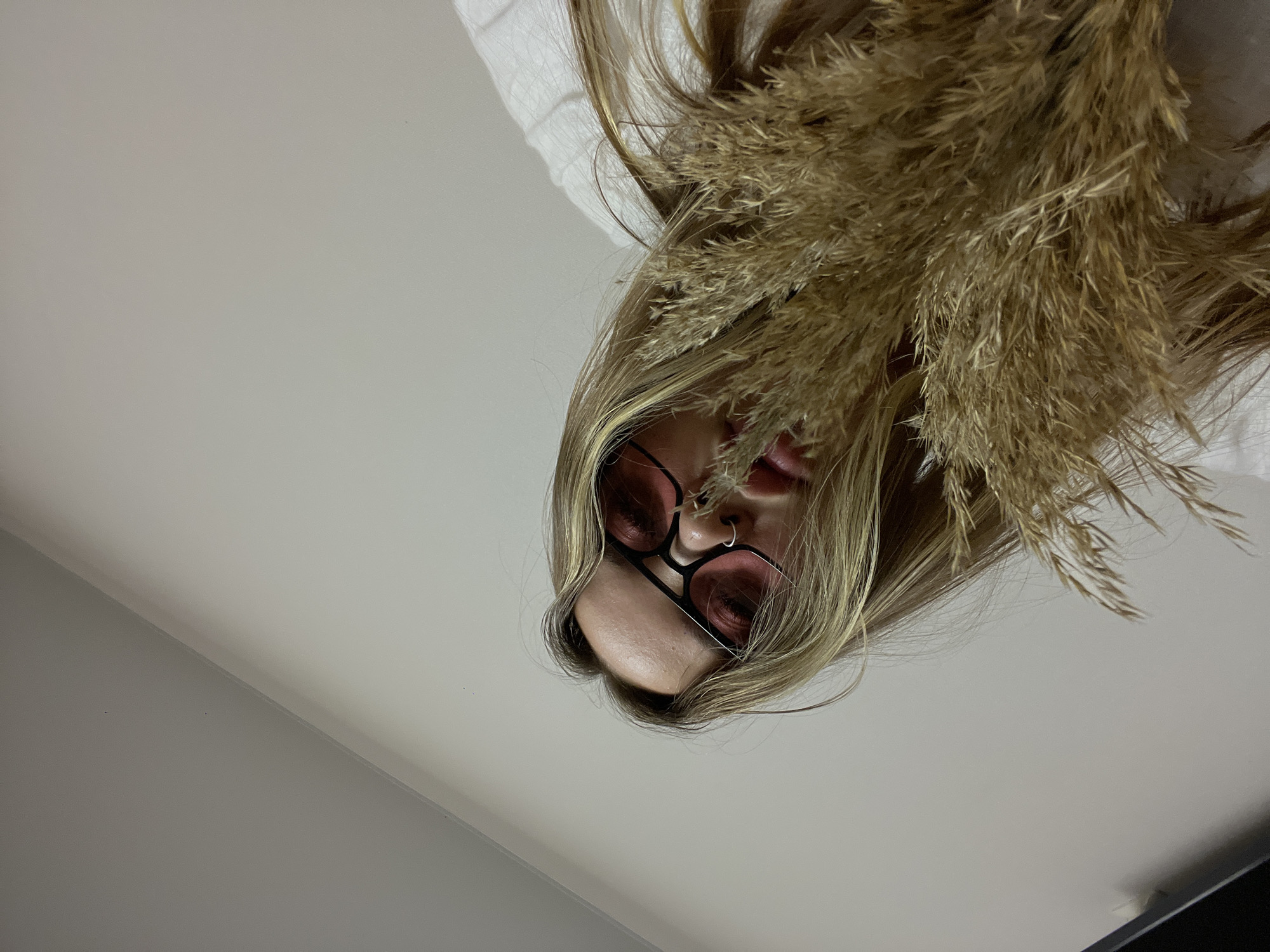 The image features a close-up, angled shot of a person, likely female, captured from a low perspective looking upwards (or downwards if the image is rotated to be upright). The subject's face is partially visible, framed by long, light brown or blonde hair. She wears distinctive round-framed glasses with reddish-pink tinted lenses and a small silver hoop nose ring on her left nostril. Her lips are slightly parted, revealing a hint of pink.
A prominent feature of the image is a large arrangement of dried, feathery pampas grass or similar plant material intertwined with and obscuring a significant portion of her hair, particularly on the right side of the frame. The grass is a warm, golden-brown color, creating a striking contrast with her hair.
The background is minimalist and plain, consisting of a light grey or white wall or ceiling, with subtle variations in shade suggesting a corner or natural light fall-off. There are no other discernible objects or details in the background. The scene suggests an indoor setting, likely a styled portrait or artistic shot. No specific activity is taking place beyond posing for the photograph. There is no visible text. The location context for this image is Krasnodar, Russia.