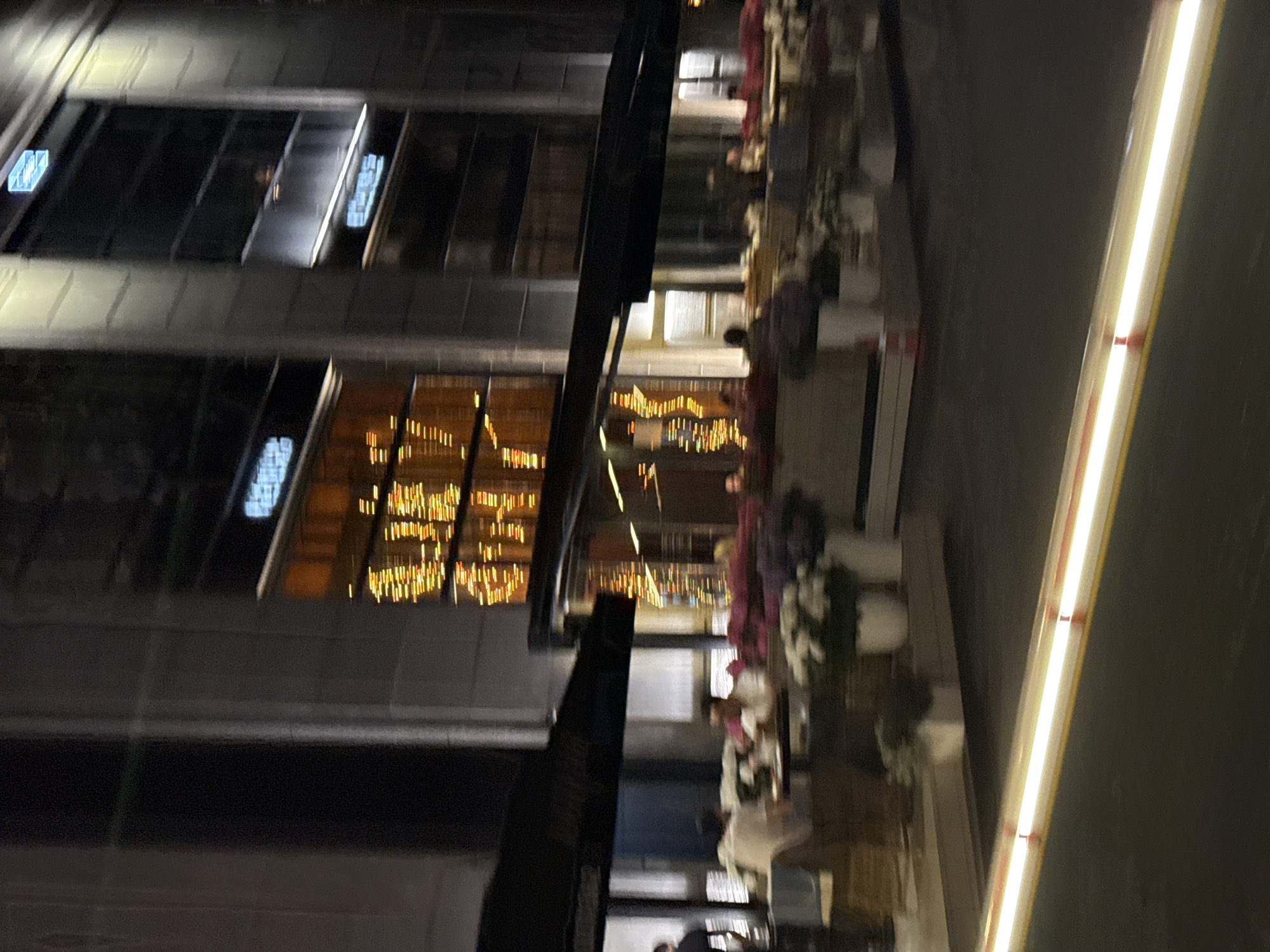 The image depicts a nighttime urban scene in Moscow, Russia, focusing on the exterior of a large commercial building, specifically identified by the prominent "ЦУМ" (TSUM) signage visible in its upper windows.
The building, occupying the background, is multi-story with a dark, possibly stone or glass, facade featuring numerous large windows. Many windows display vertical patterns of bright, warm-toned decorative string lights. In the upper-left section, a glowing blue sign with the Cyrillic letters "ЦУМ" is clearly visible within a window, indicating the renowned department store. Other indistinct blue-lit signs are present in adjacent upper windows.
In the middle ground, at street level, an outdoor cafe or restaurant operates beneath several black awnings. Numerous tables and chairs are arranged on a light-colored paved area. Multiple individuals, estimated between 15-25, are present, seated and standing, though their features are blurred due to motion and low light. They appear to be patrons dining or socializing. Potted plants with white and pink/purple flowers are distributed throughout the outdoor seating area, resting on the ground and on low barriers.
The foreground consists of a dark asphalt surface, presumably a street or sidewalk. A bright, elongated horizontal light streak, indicative of a moving light source (such as vehicle headlights/taillights or an illuminated barrier captured with a slow shutter speed), traverses the lower portion of the frame from left to right, angled slightly upwards. This light trail contains faint, regularly spaced vertical red segments within its glow.