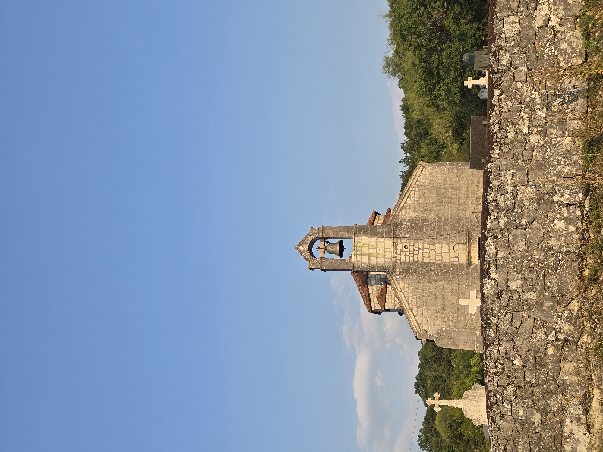 This is a vertical, medium-shot, outdoor, daytime photograph of a small stone church in Medun, Montenegro. The church has a bell tower with a bell. In front of the church is a stone wall, and behind the wall, within the church grounds, are several gravestones and crosses. The sky is clear and blue, with a few wispy clouds. The church is made of rough-hewn stone, and its roof is tiled. The bell tower is on the front of the church. The gravestones vary in size and material, and some are topped with crosses. The environment is rural, with trees visible in the background. The lighting suggests it is late afternoon.