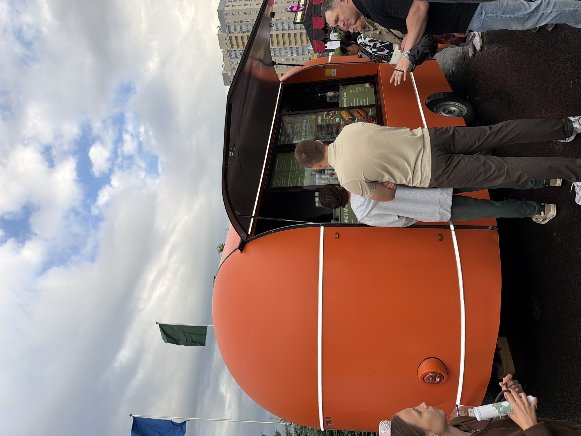 In this eye-level, outdoor shot, an orange food truck is the central focus, with its service window open. Several people are gathered around it, seemingly waiting to order or receive food. The truck is parked on asphalt, and a backdrop of buildings and a cloudy sky suggests an urban or suburban setting.
The food truck itself has a distinctive, rounded design, painted in a bright orange hue. A white stripe encircles the lower portion of the truck. The open service window reveals a menu board displaying various food items, including "КОТ-ДОГИ" (hot dogs) and "ПАНІННИКИ" (pancakes), with prices listed. A few bottles and containers are visible on a shelf inside the window.
To the left of the truck, a woman in a brown hoodie is holding a white cylindrical object. She is looking towards the truck, her body angled in that direction. Another person, wearing a grey hooded sweatshirt and dark pants, stands with their back to the camera, facing the service window.
In front of the service window, a man in a light brown polo shirt and dark pants also stands with his back to the camera. To his right, a group of people are conversing. Among them is a woman in a black and white patterned top and a beige jacket, holding a white bag. Next to her, a man in a black t-shirt and jeans gestures with his hand, appearing to be in conversation. Further to the right, another woman is partially visible.
Two flags are planted on poles to the left of the food truck, one blue and one green. The sky above is filled with dramatic, gray clouds interspersed with patches of blue. The overall atmosphere suggests a casual outdoor gathering or event.