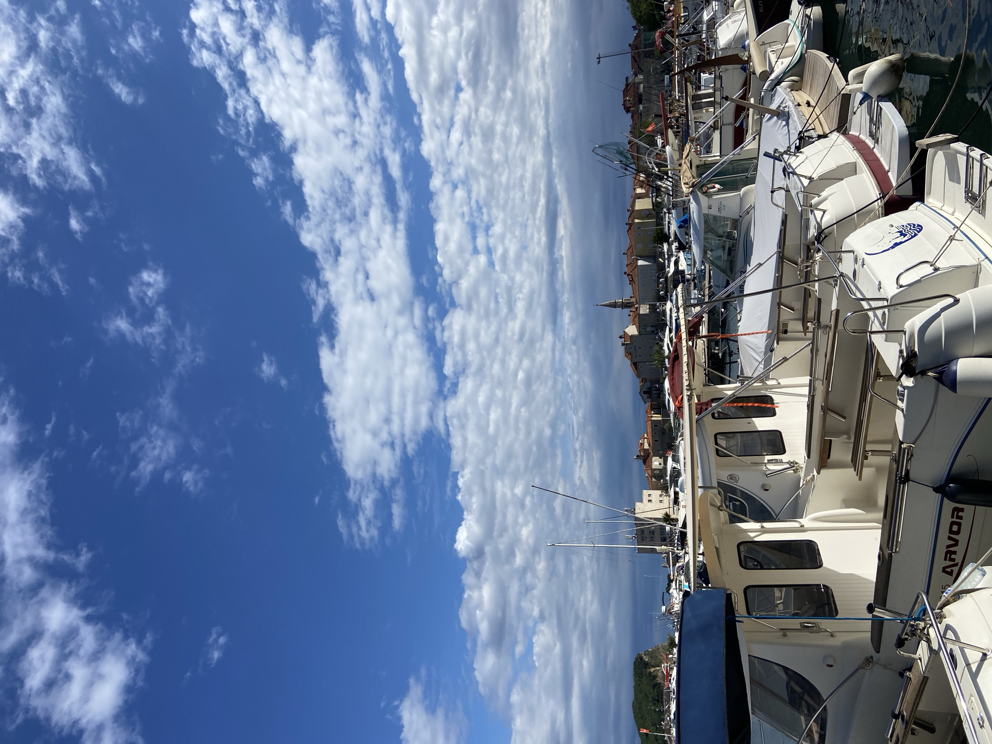 This full-color, eye-level photograph captures a vibrant marina scene under a bright, partly cloudy sky. The image is taken from the water, looking towards a bustling coastal town.
The primary subjects are numerous white leisure boats and yachts of varying sizes, densely packed and docked side-by-side in calm water. Many boats feature protective covers, shiny chrome railings, and exposed decks. Visible details on the boats include cylindrical fenders hanging along their sides, masts, and rigging. One boat clearly displays the brand name "ARVOR" in red lettering on its hull, while another features a distinctive blue emblem, possibly a star or a stylized octopus, on its stern. No people are explicitly visible, suggesting a peaceful, uncrowded moment.
Behind the marina, a picturesque town with traditional Mediterranean architecture stretches along the coastline. Buildings are characterized by light-colored facades and numerous terracotta-tiled roofs. A prominent church steeple or clock tower rises above the other structures, adding a historical touch to the urban landscape. Distant hills or mountains can be seen in the far background, partially obscured by the town.
The weather appears to be excellent, indicative of a pleasant day. The sky is a vivid deep blue, particularly overhead, transitioning to a lighter hue towards the horizon. It is adorned with scattered, fluffy white cumulus clouds that stretch across the frame. The bright sunlight suggests it is daytime, likely late morning or early afternoon. The water in the marina is still and reflects the hulls of the boats and the overhead sky.
Based on the provided context, this scene is located in Budva, Montenegro, depicting its scenic harbor and coastal charm. The overall impression is one of tranquility and an inviting maritime atmosphere.