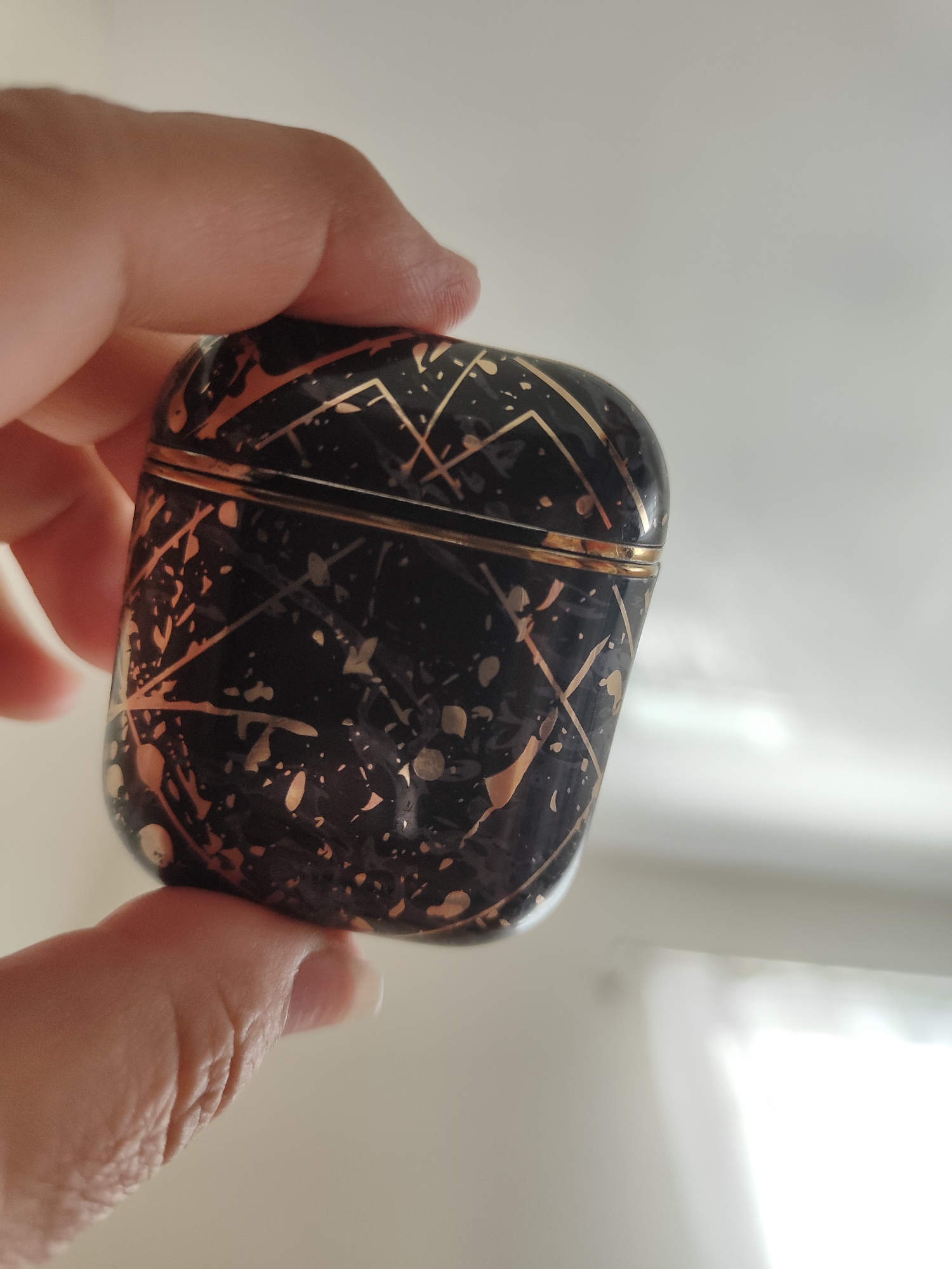 A close-up shot captures a hand holding a small, decorative black object with a marbled rose-gold pattern. The object is rounded with a distinct seam around its middle, suggesting it might be a container or case. The hand's fingers are gently cupping the object, with the thumb and index finger providing support. The surface of the object is dark, with irregular splatters and thin lines of rose-gold that create a dynamic, abstract design. The background is softly blurred, appearing to be a bright, neutral space, possibly an indoor environment with diffused light. The lighting is soft and even, highlighting the texture and sheen of the object. No text is visible in the image.