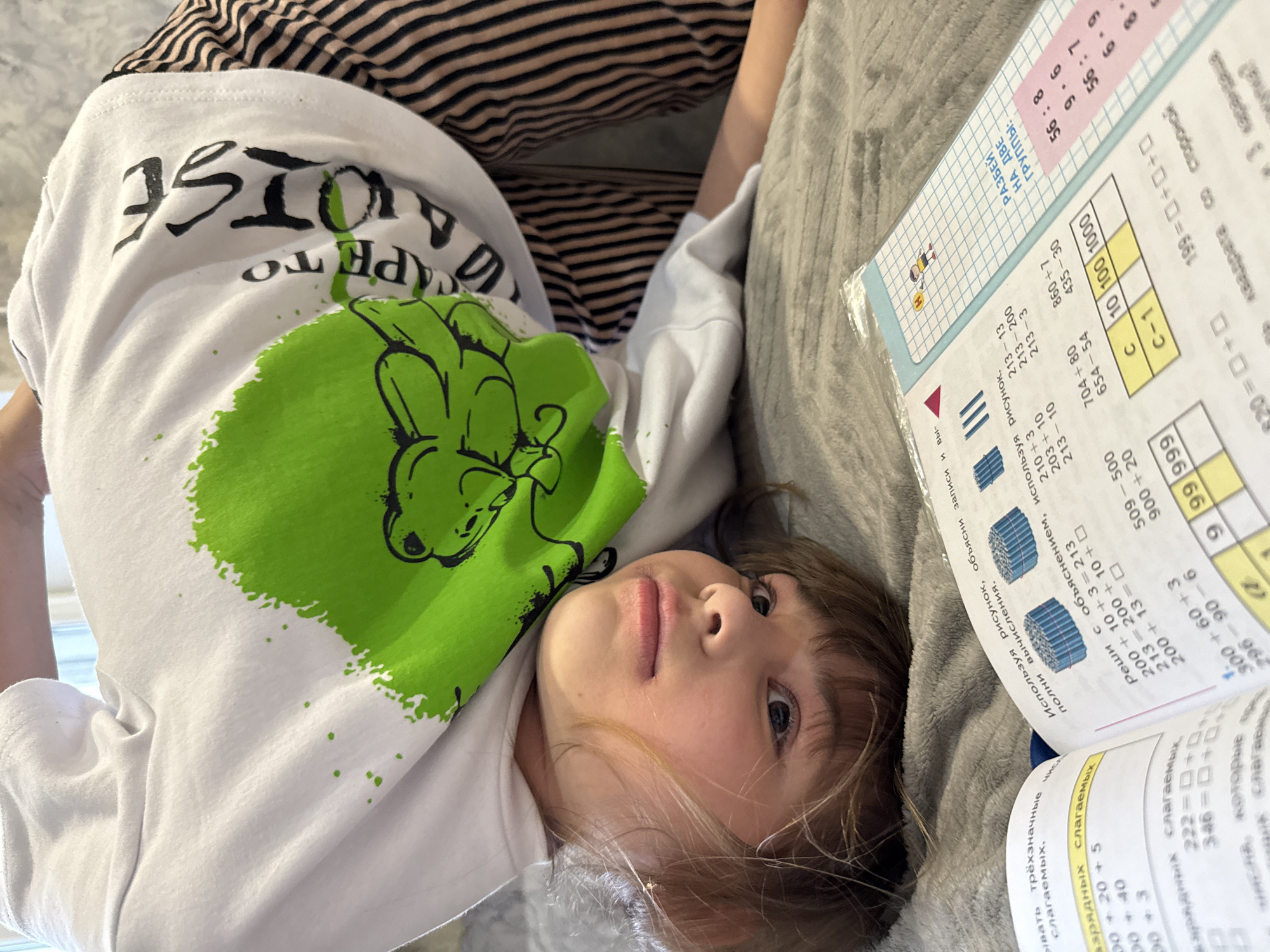 A child with shoulder-length brown hair is lying on a gray textured surface with an open math workbook. The workbook features columns of numbers and mathematical problems, some in Cyrillic script. The child is wearing a white long-sleeved shirt with a large green graphic and black text. The child is also wearing striped pants. The image shows the child looking directly into the camera. It is impossible to identify the city, Yaroslavl', based on visual information.