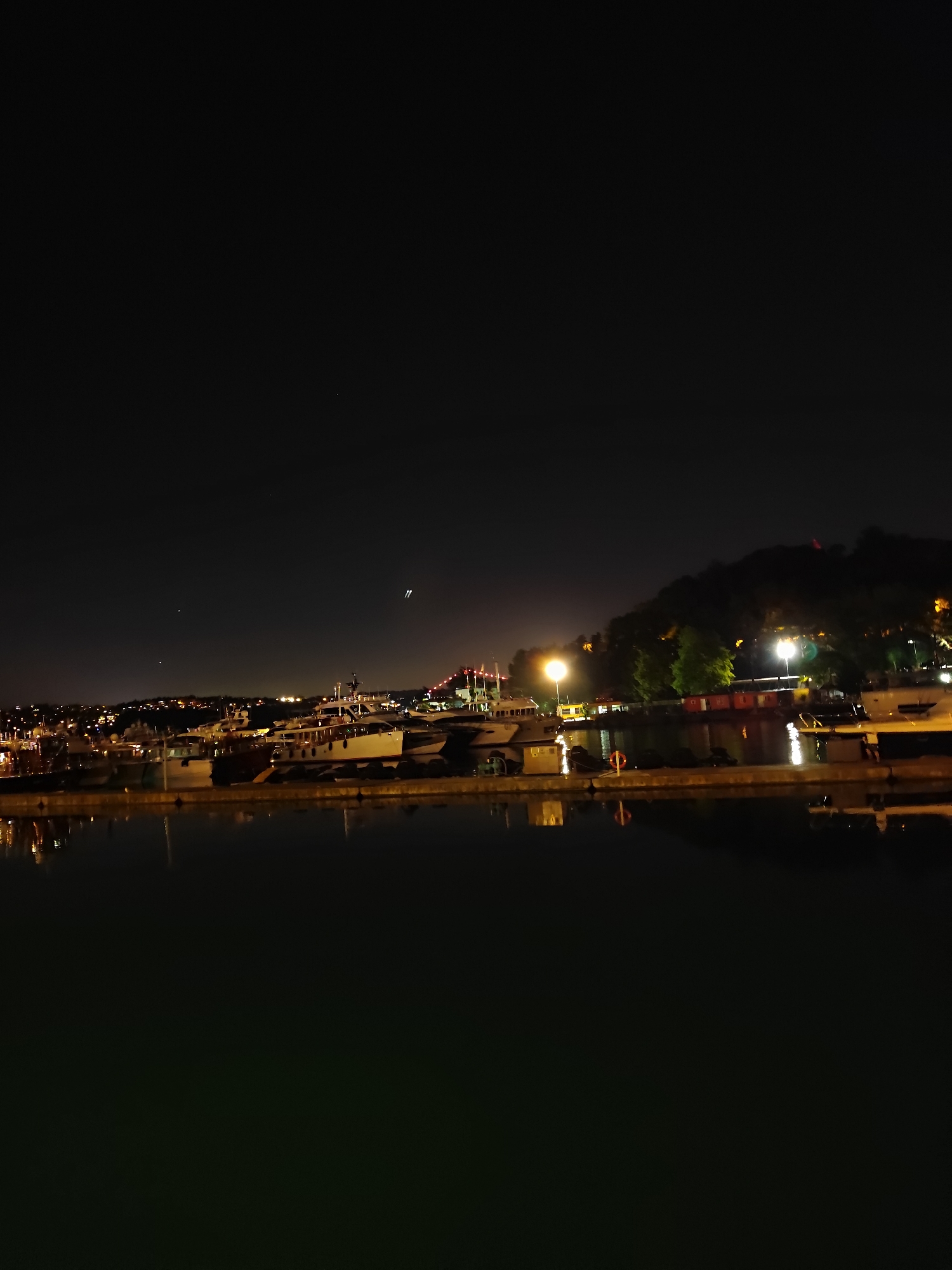 The image captures a nighttime view of a marina with several boats docked. The foreground is dominated by dark water reflecting the lights from the shore. The middle ground shows a line of boats, some of which are illuminated, and a pier. In the background, a hillside is covered in trees and dotted with lights from buildings. The sky is dark and clear, with a few faint stars visible, and what appear to be airplane contrails. The scene is likely located in a coastal urban area, potentially a marina in Sarıyer, Turkey, given the visual characteristics of a bustling waterfront at night.
