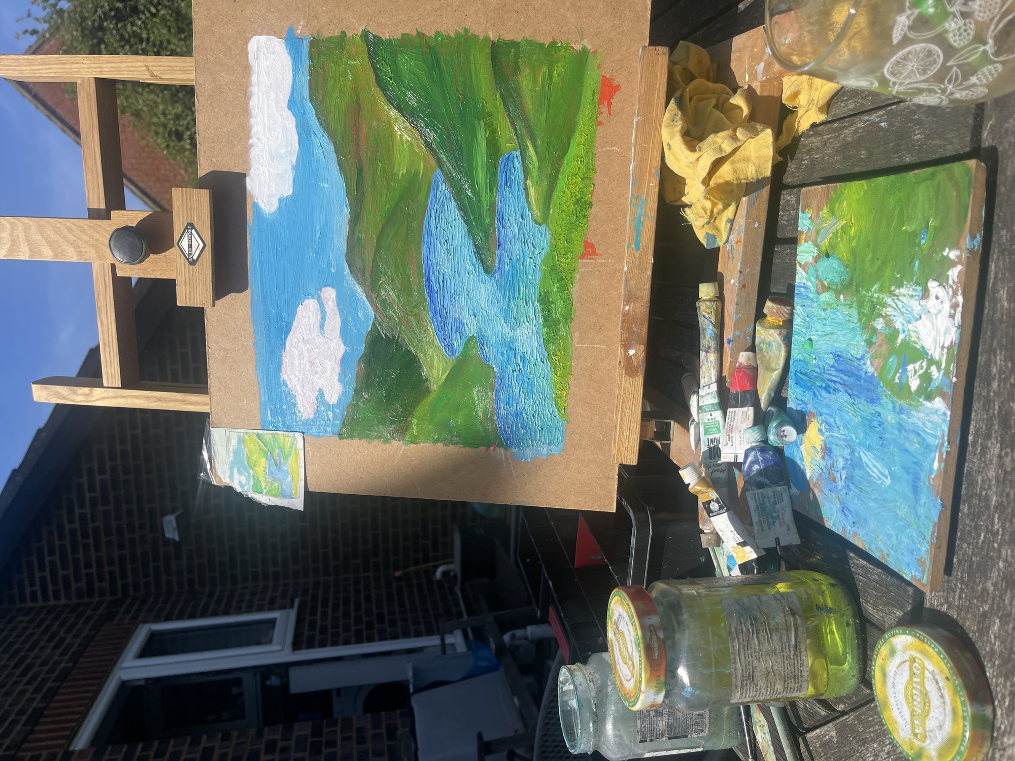 The image depicts an outdoor painting setup on a wooden table or surface. A painting of a landscape with green hills, a blue waterfall, and white clouds is mounted on a wooden easel. Beside the easel are tubes of paint, a palette with mixed paints, jars, and a yellow cloth. In the background, a brick building with a window is visible, along with a glimpse of a blue sky and greenery. The location is possibly in the city of Maidstone, United Kingdom. There are no people visible in the image.