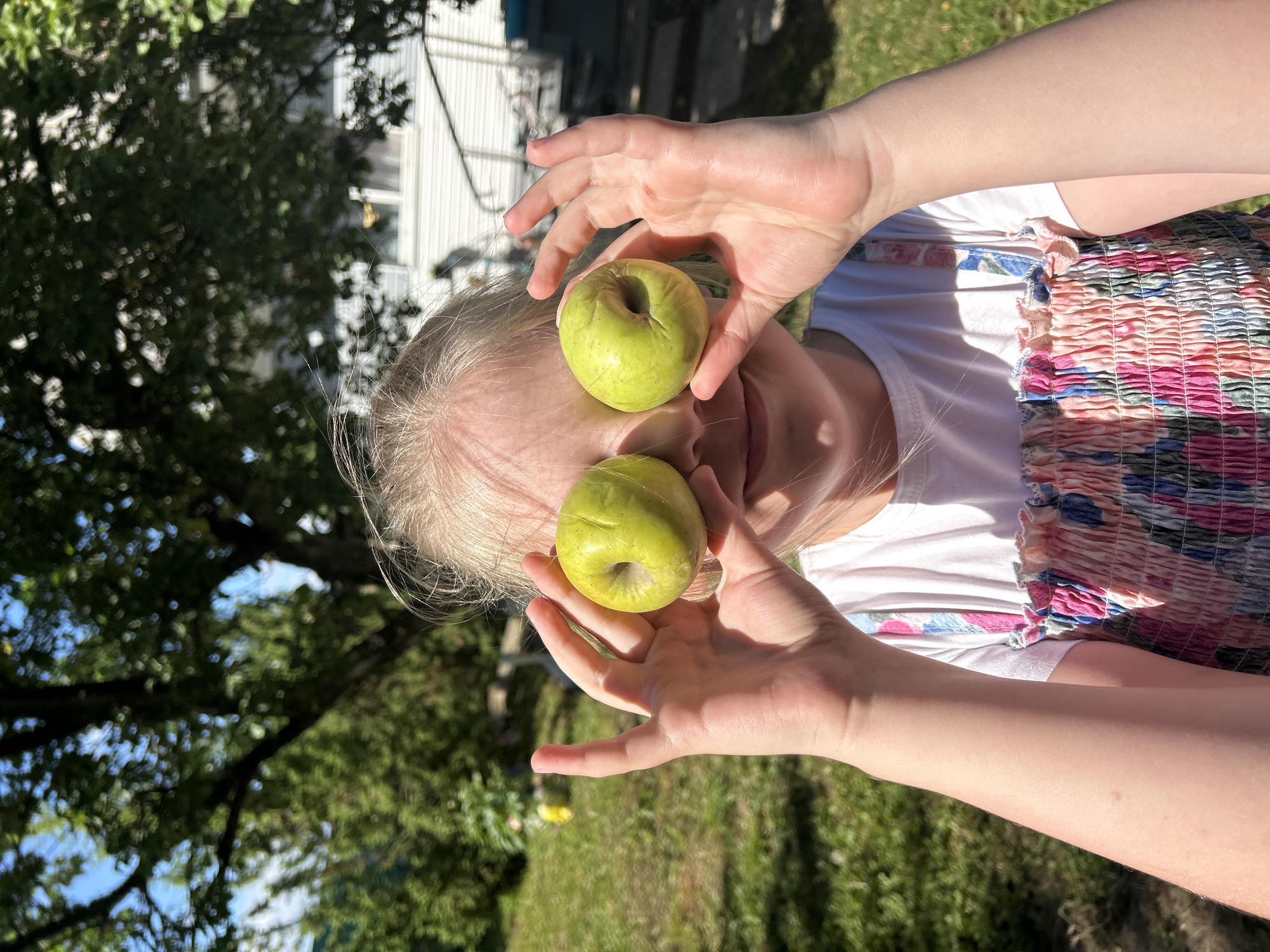A young girl with blonde hair is holding two green apples in front of her eyes outdoors. She is wearing a white t-shirt and a dress with a floral pattern. The background is a grassy yard with trees and a house. The lighting suggests it is daytime. The image is a close-up shot of the girl's face and upper body.