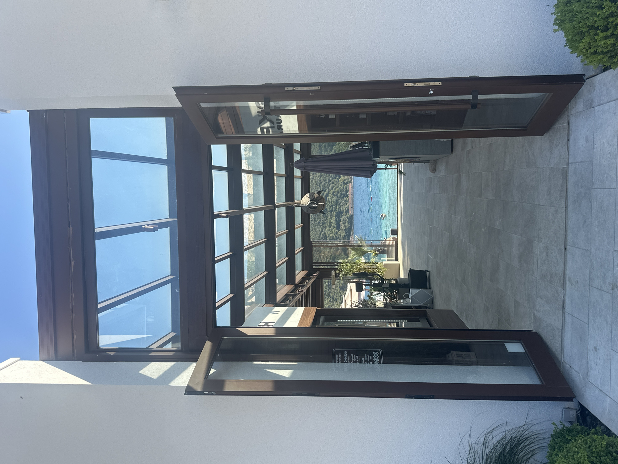 The image shows an open doorway leading to a covered patio overlooking a body of water. The patio has a glass roof with wooden beams and is paved with large, light-colored tiles. Beyond the patio, there is a view of the sea with a few boats and a person swimming. The surrounding hills are covered in lush green vegetation.
The architecture appears modern and elegant, with white stucco walls and dark wooden door frames. Inside the entrance, to the left, a glass-paned door is partially ajar, revealing a sign with text that is difficult to read but appears to list business hours. To the right, another door is also open, showing shelving with various items. On the patio, there are potted plants and a folded umbrella. The lighting suggests it is daytime, with clear blue skies overhead. The overall scene conveys a sense of tranquility and a connection with the natural environment. This location is likely within the vicinity of Kumbor, Montenegro, known for its coastal beauty and resorts.