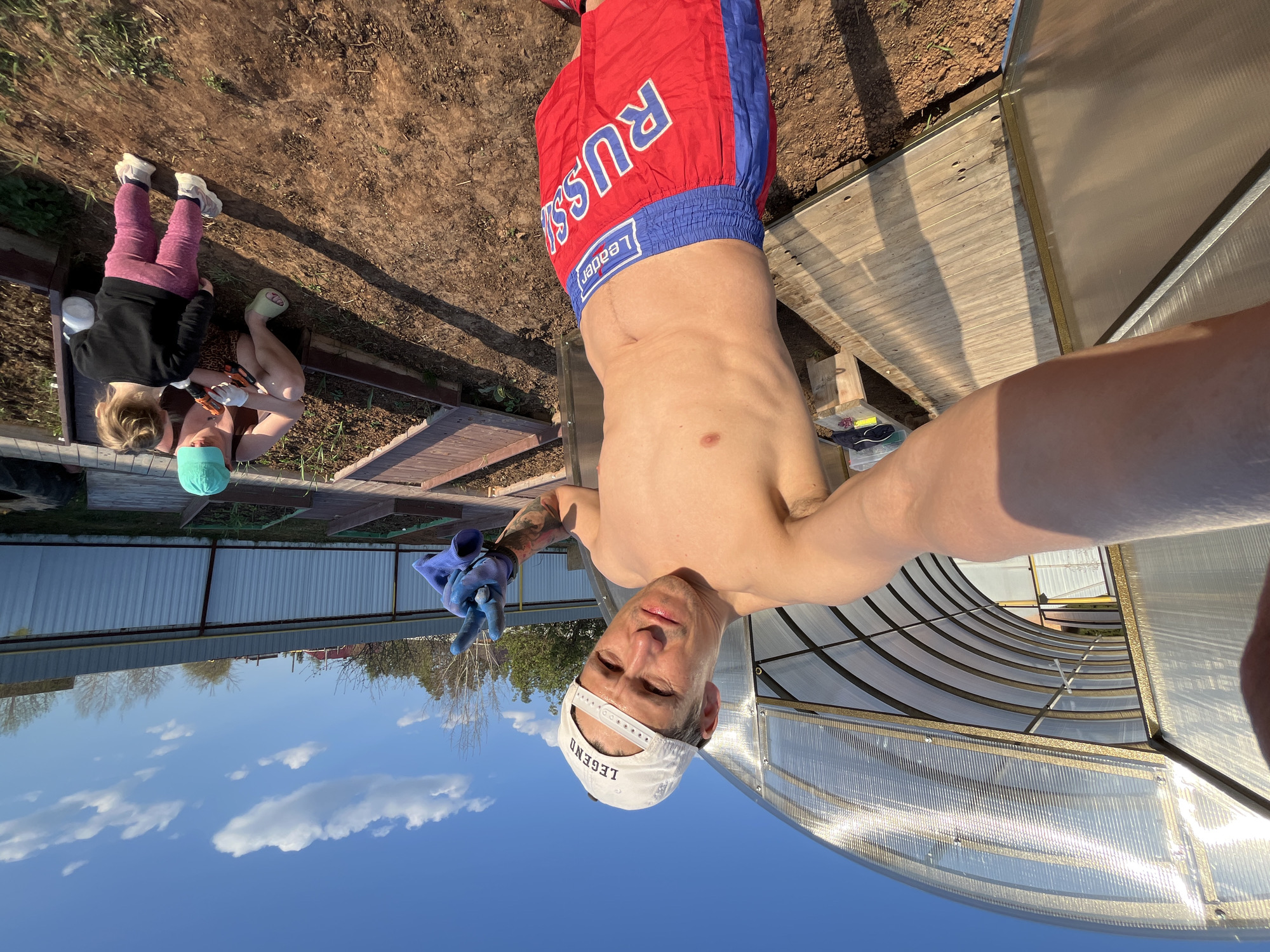 A man in a white cap with the word "LEGEND" on it, blue work gloves, and red and blue boxing shorts labeled "RUSSIA" is shown in a selfie. He is shirtless and making a peace sign with his left hand. Behind him, a greenhouse with curved, translucent panels stands under a partly cloudy sky. In the background, two people appear to be working on a garden or yard; one is kneeling and the other is sitting. The photo was taken in Chaikovsky, Russia.