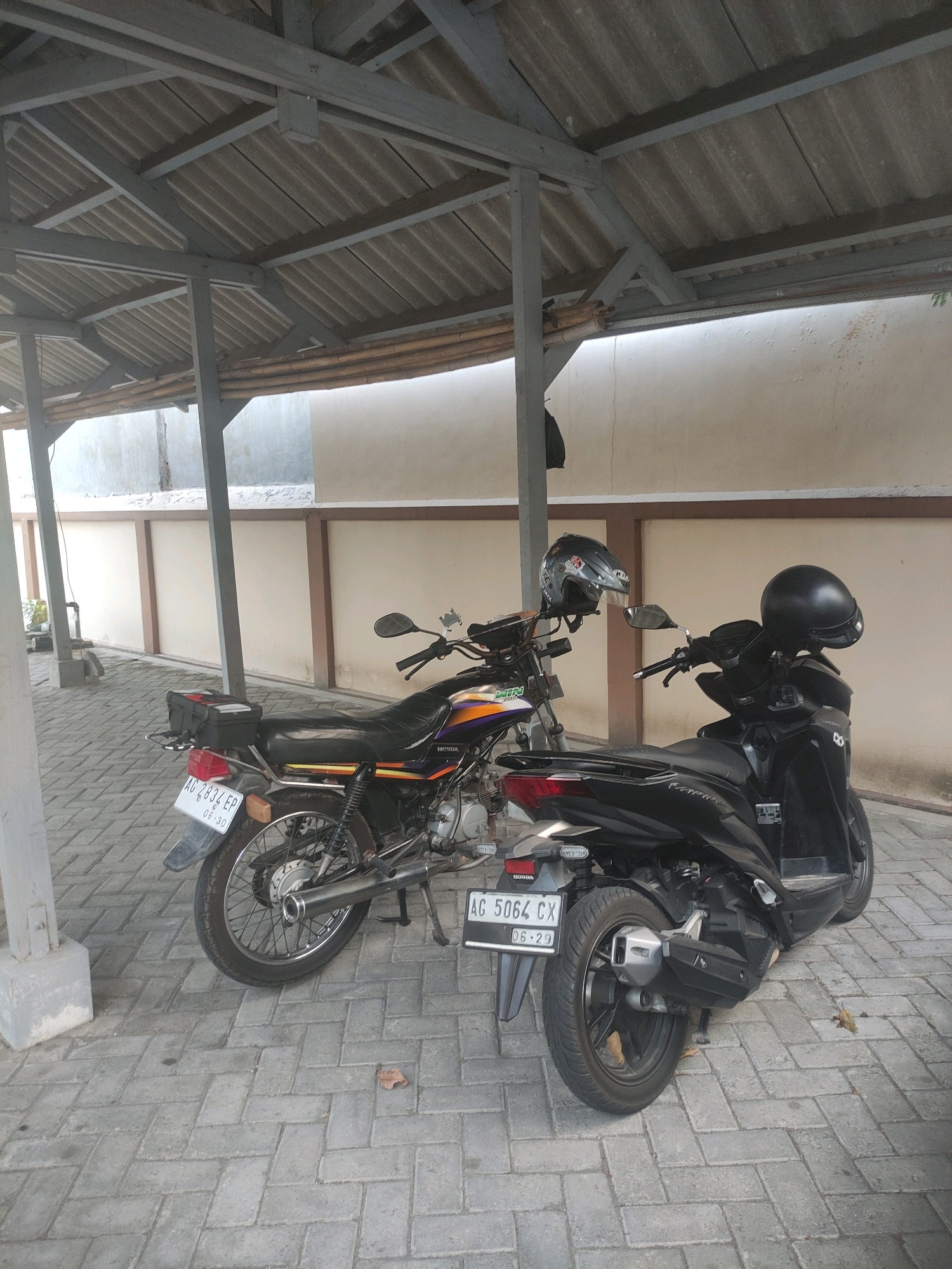 Two motorcycles are parked side-by-side under a covered structure. The motorcycle on the left is black with purple and orange accents and has a license plate reading "AG 2834 EP". It has a black storage box on the rear. A grey helmet with stickers is hanging from the handlebars. The motorcycle on the right is black and appears to be a scooter. Its license plate reads "AG 5064 CX". A black helmet is placed on the handlebars of this motorcycle. The motorcycles are parked on a paved surface made of grey interlocking bricks. The covered structure has a corrugated metal roof supported by grey metal beams and wooden poles. The background consists of a light-colored wall with some wooden trim. The scene is located outdoors in what appears to be a residential or commercial area in Ngasem, Indonesia.