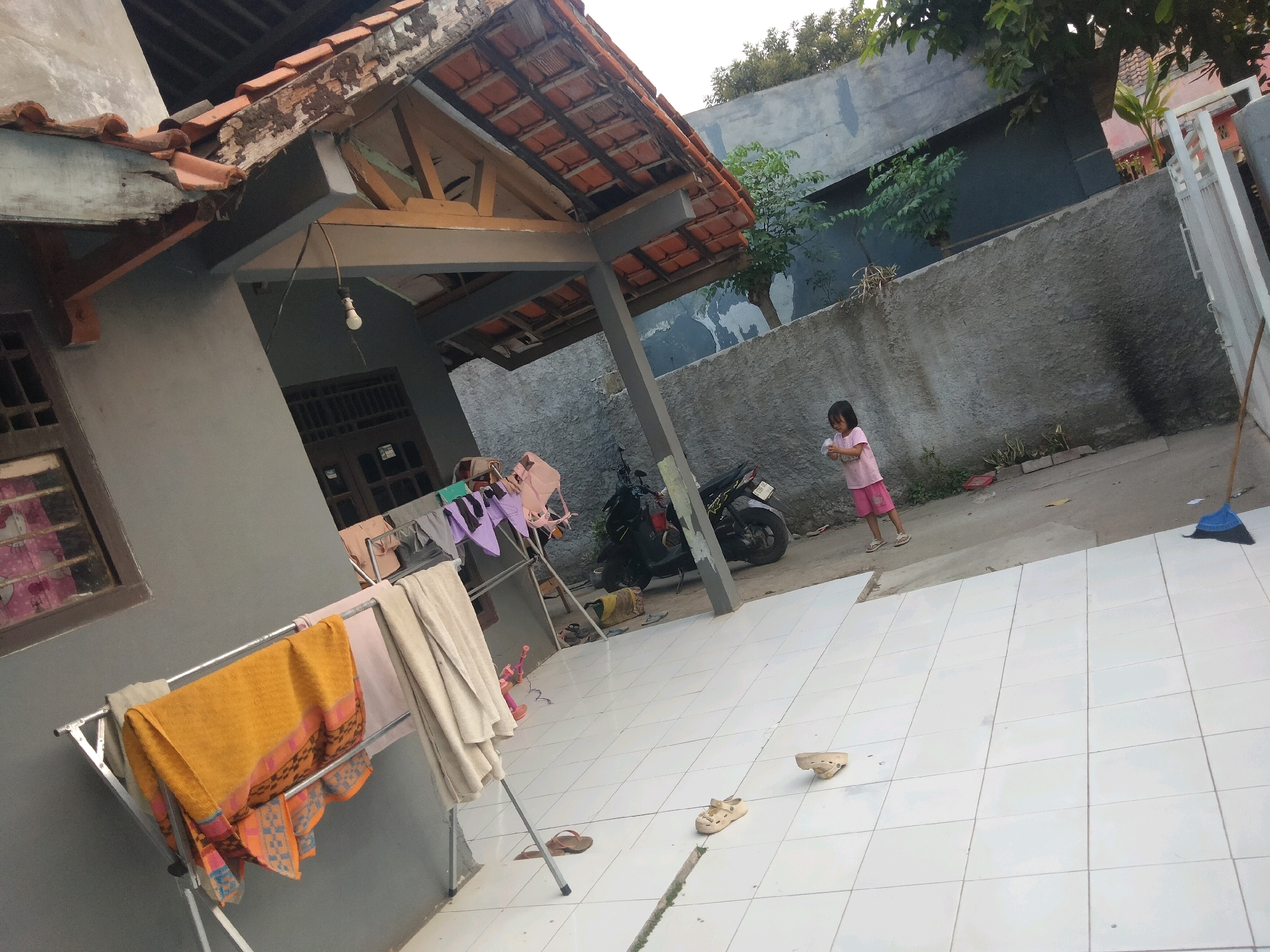 In the city of Suka Sari, Indonesia, a residential outdoor scene is depicted. A young girl in a pink t-shirt and shorts stands in the middle of a tiled courtyard, holding a white object. To her left, a parked motorcycle is visible. In the foreground, a clothes drying rack holds various items of laundry, including a vibrant orange cloth with a patterned border and lighter-colored towels. A grey wall forms the backdrop, with a section of a tiled roof and wooden beams of a building on the left. A blue broom with a wooden handle leans against the wall on the right. Several pairs of sandals are scattered on the tiled ground. The environment appears to be a domestic backyard or patio area.