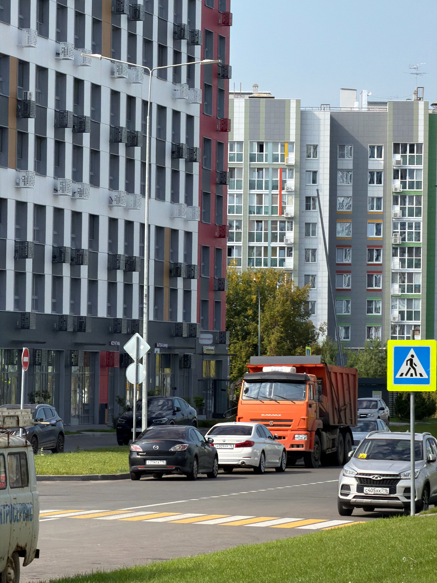 In an urban environment, a busy street scene unfolds under a clear sky. A pedestrian crossing is marked with yellow and white stripes on the asphalt. Several cars are visible: a silver Chery SUV in the foreground with license plate C405XK 716, a black Mazda sedan with license plate T714TA 716, a white BMW sedan, and a dark-colored SUV to the left. In the background, an orange dump truck is parked, and behind it, tall, modern apartment buildings with colorful architectural details dominate the skyline. A pedestrian crossing sign with a blue background and a white silhouette of a person walking is posted on a pole. Streetlights are present, and some trees add greenery to the scene. The buildings feature numerous balconies with railings and air conditioning units.