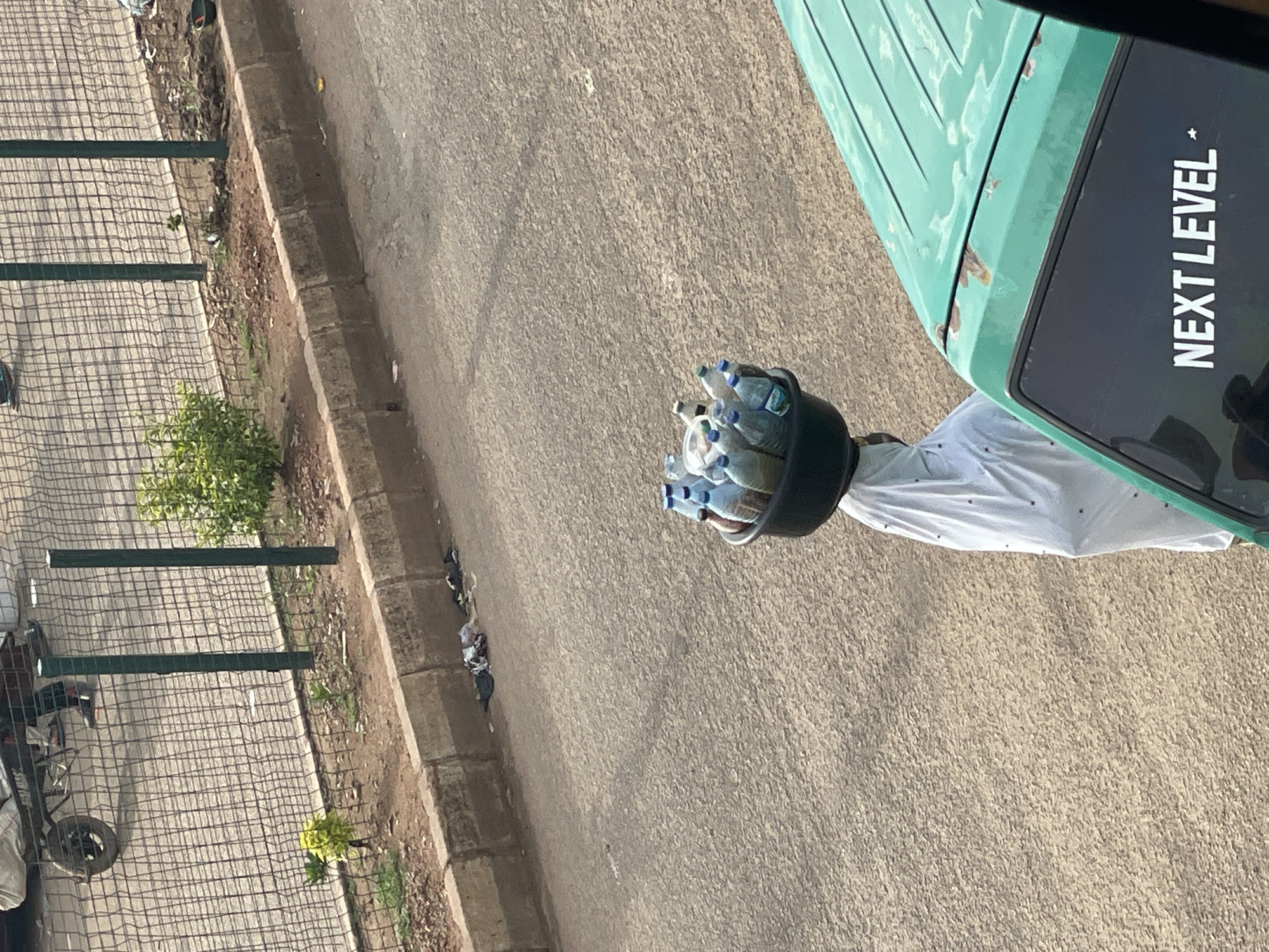 A person is seen from above, carrying a bucket filled with plastic bottles. The person is wearing a white garment. The bucket is black and appears to be made of plastic or metal. The bottles are clear plastic and are filled with a clear liquid, likely water. They are arranged in a dense cluster within the bucket.
The scene is outdoors, on a paved surface that appears to be a road or sidewalk. To the left, there is a metal fence with a diamond-shaped mesh pattern, and beyond that, a patch of dry dirt with some sparse vegetation. A small, green bush is visible near the fence. Further to the left, some objects, possibly part of a vehicle or cart, are partially visible.
To the right of the person, a portion of a green vehicle is visible. On the side of this vehicle, there is a black rectangular panel with white text that reads "NEXT LEVEL". The text is capitalized and has a star symbol next to it. The vehicle appears to be a van or truck, and the green surface shows some signs of rust or wear.
The overall lighting suggests it is daytime, likely sunny, as indicated by the shadows and the brightness of the scene. The ground texture is rough and granular. There are some scattered pieces of litter on the edge of the paved area near the fence. The image is oriented in a way that the subjects and the vehicle are tilted, creating an unusual perspective. The focus is primarily on the person and the bucket of bottles, and the side of the green vehicle.