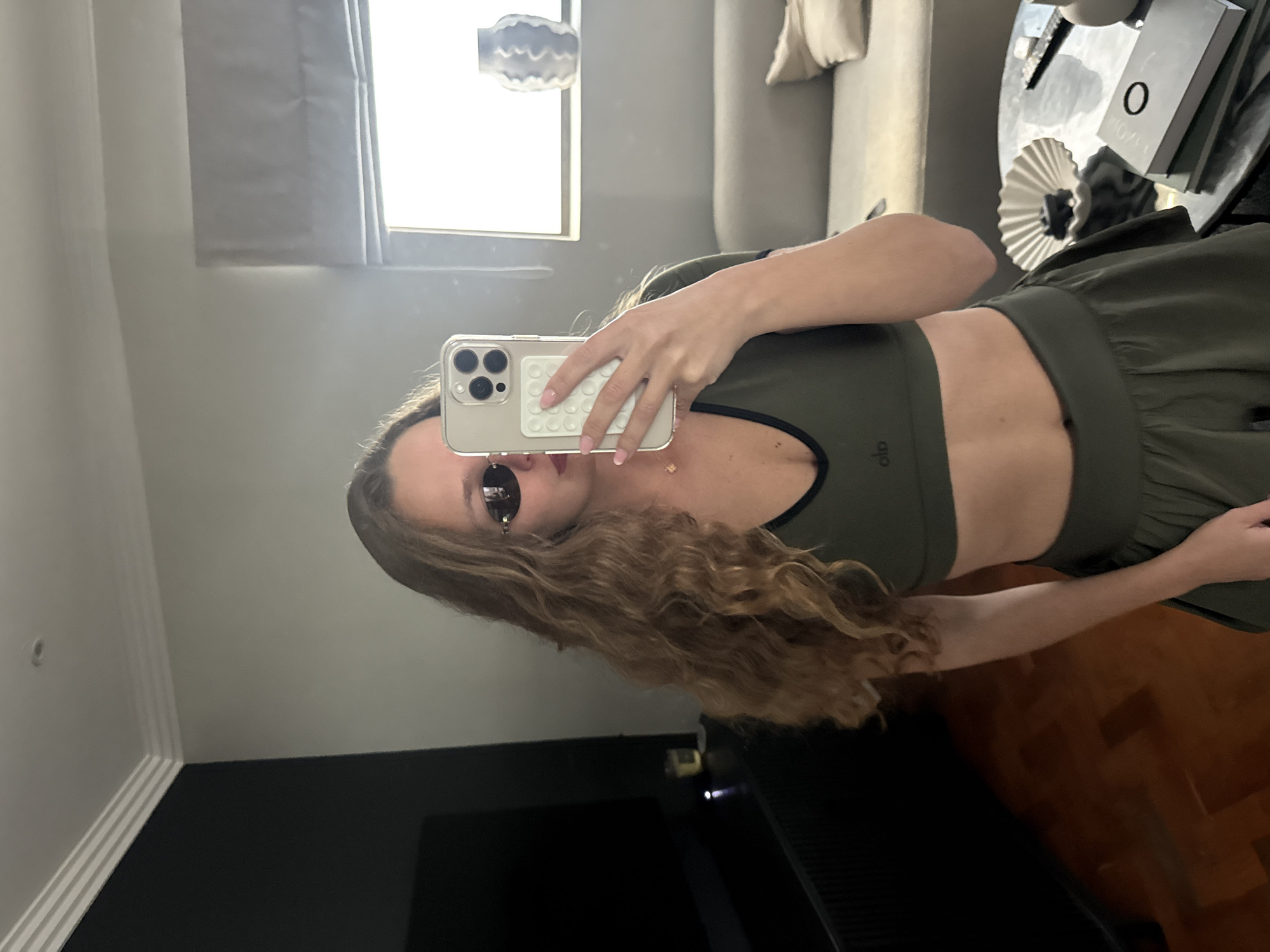 A woman stands indoors, presumably in a room or apartment. She holds a cell phone up to take a selfie in a mirror. The woman wears a matching olive green athletic crop top and skirt or shorts. She has long, curly brown hair and is wearing sunglasses. Reflected in the mirror is a window with a view of the outside, a couch with pillows, and a side table with objects on it. The area underneath the mirror appears to be a dark, possibly black, surface. There is no evidence of Cape Town, South Africa.