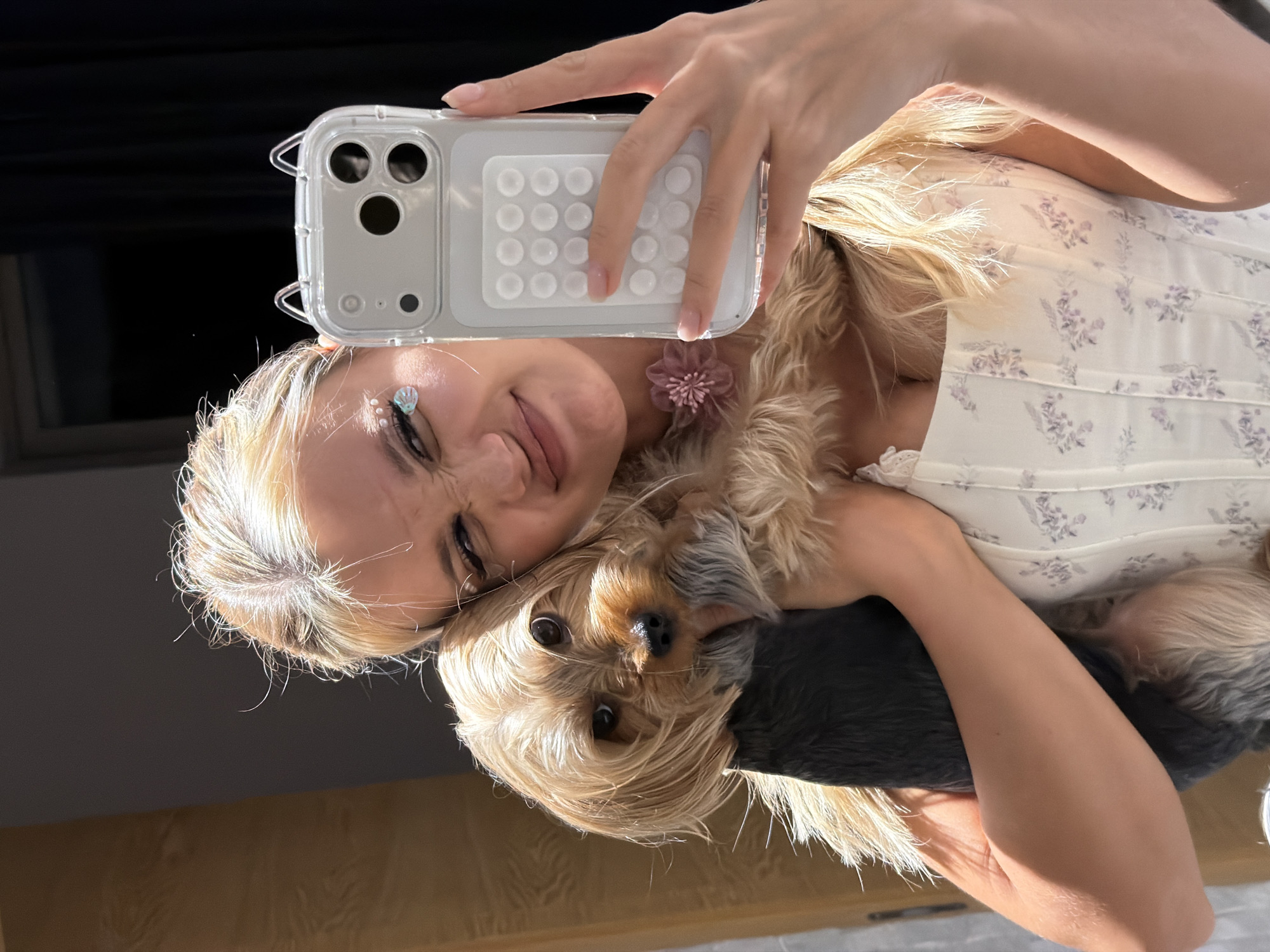 A young Caucasian woman with blonde hair is taking a selfie with her phone, which is held in her left hand. She is embracing a small Yorkshire Terrier. The woman is wearing a white floral print dress and has light makeup with some glitter accents on her face. A small pink flower adornment is attached to her hair. The dog is peeking over her shoulder, looking directly at the camera with its ears perked up. The background appears to be indoors with natural light casting a warm glow on the subjects, suggesting it might be daytime. The setting is likely a home environment. The woman's expression is playful and happy, as she squints her eyes slightly and smiles. The dog's expression is curious.