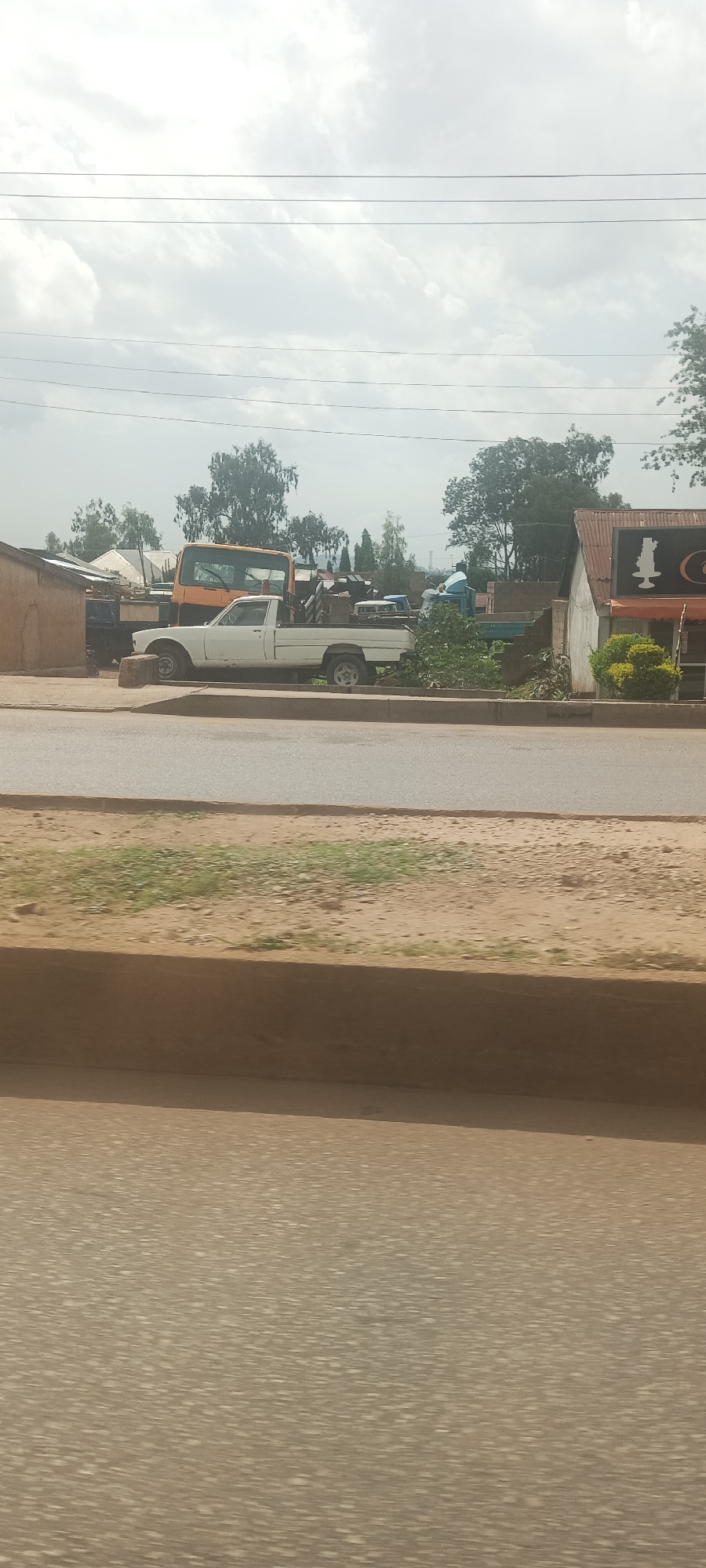 In Bukuru, Nigeria, a white pickup truck is parked on a dirt shoulder adjacent to a paved road. The truck is situated behind a low, brown retaining wall. In the background, an orange truck is visible, along with several trees and buildings. To the right of the pickup truck, a storefront with a corrugated metal roof and an awning displays a chess piece logo. The sky is overcast with some clouds.
