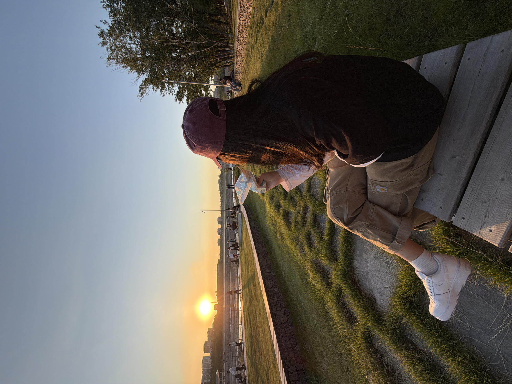 A person sits on a wooden bench at sunset, likely in Almaty, Kazakhstan, based on environmental cues. The individual wears a dark-colored shirt, tan pants, white socks, and white sneakers, and a burgundy cap. They hold a paper object, perhaps a map or snack packaging. The background includes a grassy area, a concrete barrier, a waterfront with cyclists and pedestrians, and buildings silhouetted against the setting sun.