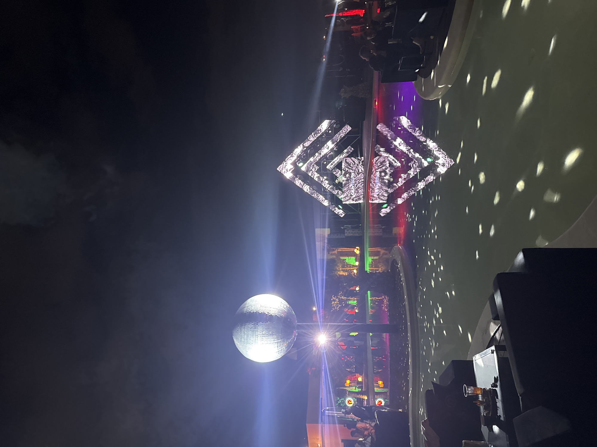 The image depicts a nighttime scene in Parakar, Armenia, showcasing an outdoor event. A large disco ball on a pole is illuminated, casting light rays. In the background, a geometric structure with light patterns is visible. A body of water reflects the lights, and a crowd of people can be seen in the distance, partially obscured by lighting effects. Closer to the foreground, there's outdoor seating, including chairs and a table with drinks, suggesting a social gathering.