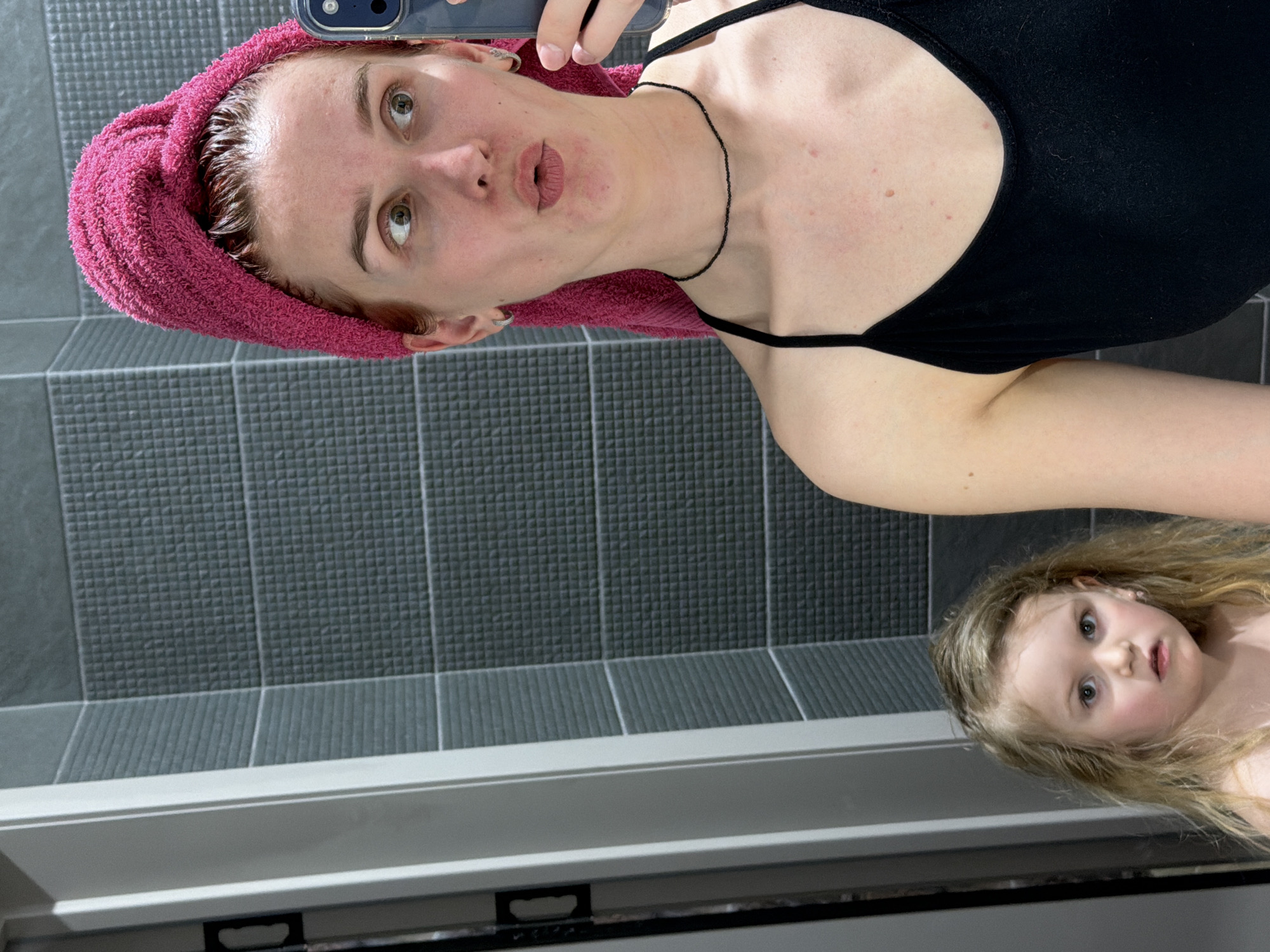 A woman with a pink towel wrapped around her head is taking a selfie in a bathroom. She is wearing a black tank top and a black necklace. A child is visible in the mirror's reflection, with blonde hair. The bathroom has gray tiled walls. The photo was likely taken in Chaikovsky, Russia.