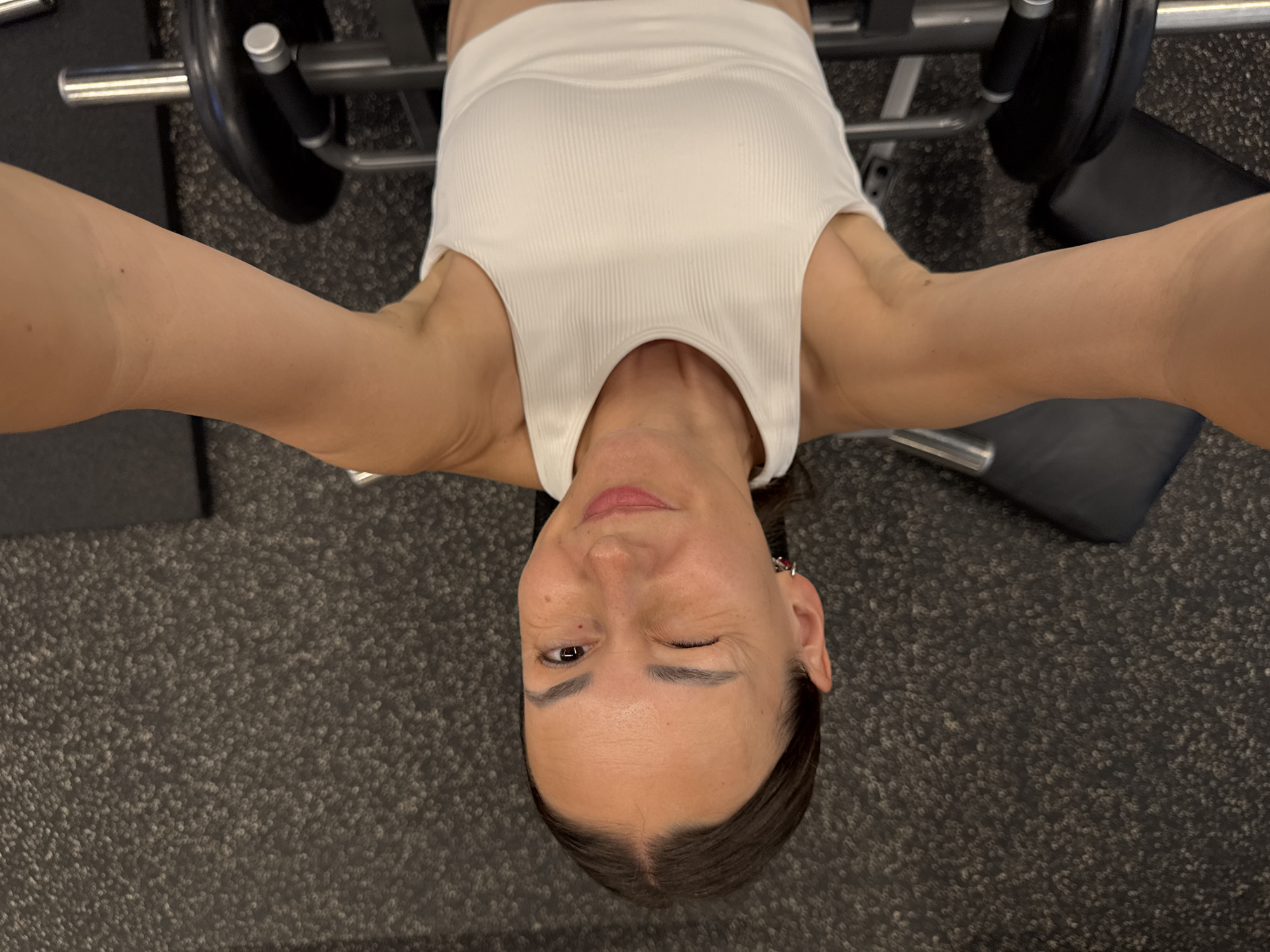 A high-angle, over-the-head shot captures a woman lying on her back on a weight bench in a gym. She is wearing a white ribbed tank top. Her arms are extended outwards, suggesting she may be in the middle of a bench press exercise or resting after one. The weight rack and a weight plate are visible behind her. The floor is covered in a textured, dark grey carpet, typical of a gym setting.
The woman is looking towards the camera and winking, conveying a playful or confident emotion. Her face is illuminated by artificial gym lighting. There are no visible indicators of the time of day or weather. The location is identified as Sochi, Russia. There is no visible text in the image.
