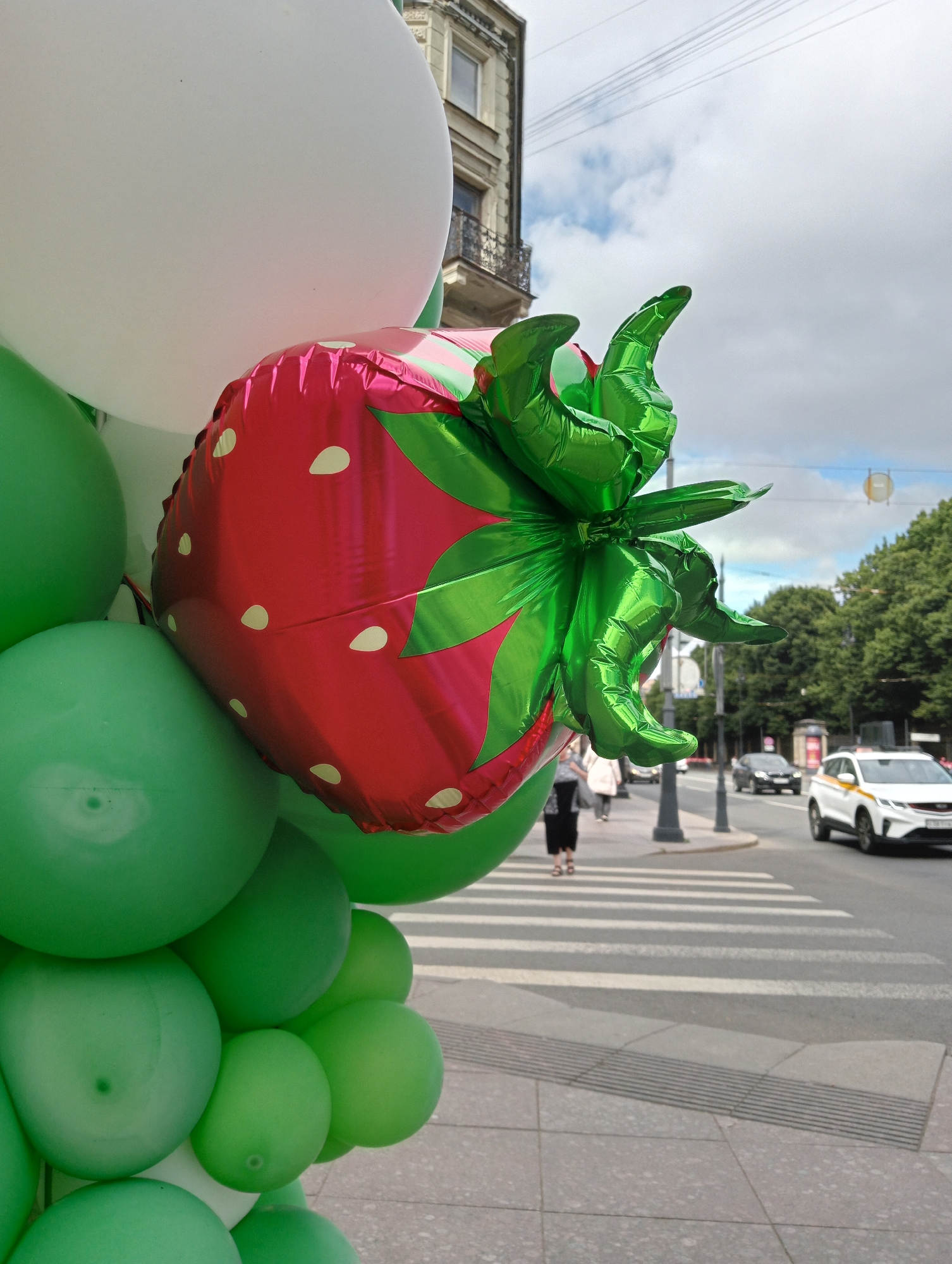 A close-up shot captures a cluster of balloons, prominently featuring a large, shiny red foil balloon shaped like a strawberry. The strawberry balloon is adorned with white dots and has metallic green foil leaves and stem. It is surrounded by numerous round green balloons and at least one large white balloon. The balloons are positioned on a sidewalk in front of a crosswalk on a city street. In the background, a multistory building with classical architecture is visible, along with trees lining the street and a few vehicles. A white taxi with black and yellow accents is in the right lane of traffic, and pedestrians are walking on the sidewalk to the left of the crosswalk. The sky is partly cloudy. The scene appears to be a street in Saint Petersburg, Russia, given the architectural style of the building and the presence of a taxi that resembles those operating in the city.