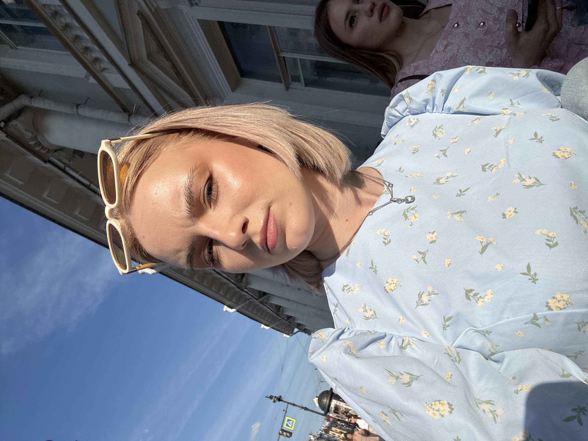The image depicts an outdoor urban scene in Saint Petersburg, Russia, on a sunny day. A young blonde female is prominent in the foreground, looking directly at the viewer with a neutral expression. She has short, light-colored hair and wears cream-colored sunglasses with amber lenses pushed up onto her head. She is attired in a light blue dress featuring a white and yellow floral pattern and a silver chain necklace with a small pendant. Sunlight illuminates the left side of her face, creating a strong highlight on her cheekbone.
To her right and partially obscured, another young female with long brown hair is visible, oriented towards the right side of the frame. She wears a pink patterned garment, and her right hand, holding a dark object, possibly a mobile phone, is visible.
The background consists of a clear blue sky with minimal white clouds and classical European architecture. A large, light-colored building with columns, cornices, and windows is situated behind the main subjects, exhibiting areas of shadow. To the left, a street extends into the distance, featuring a black pole with a traffic light and a yellow square pedestrian crossing sign. Several indistinct figures are visible walking in the far background.