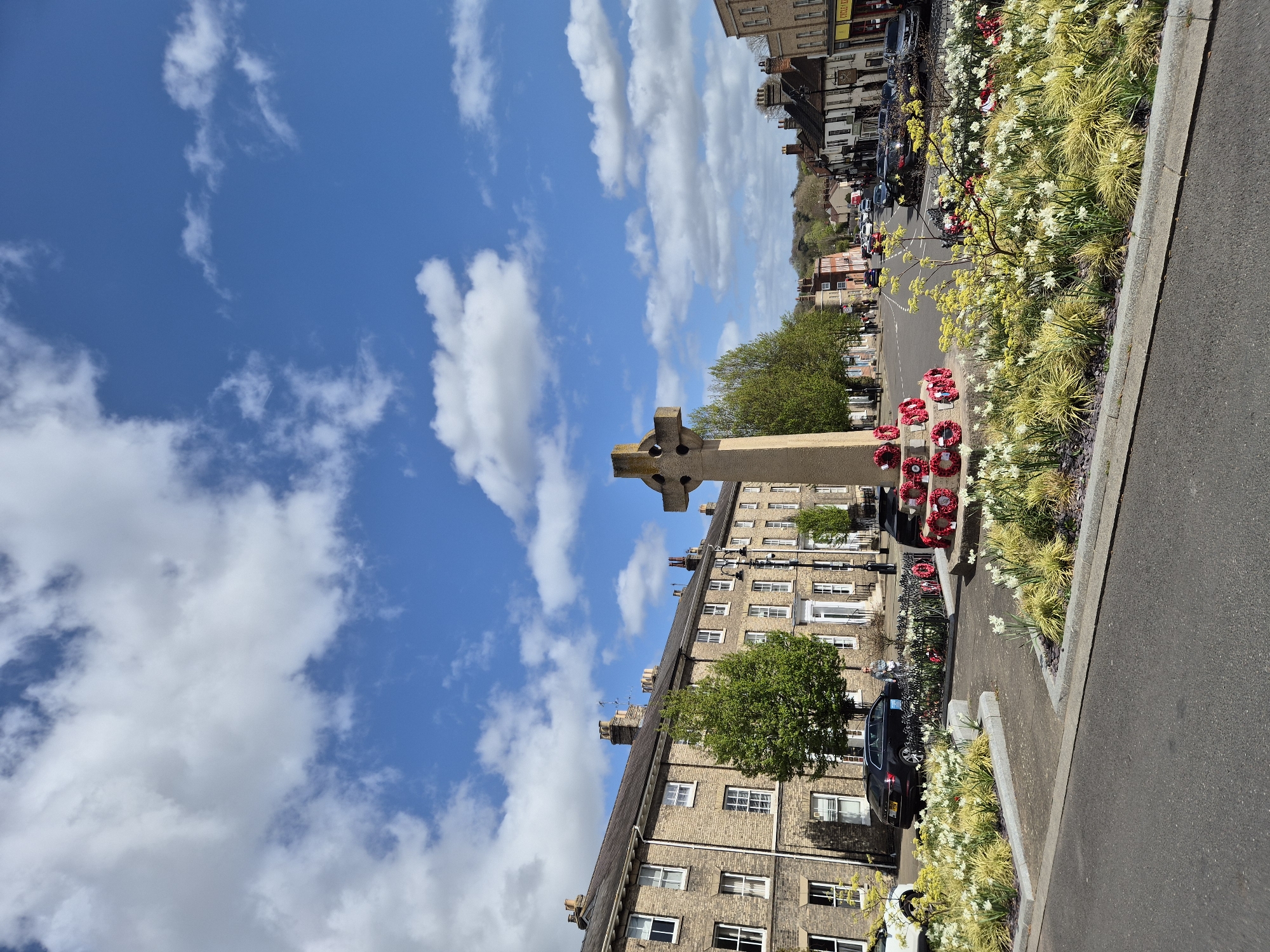 The image depicts a war memorial in Bury St Edmunds, West Suffolk, United Kingdom. The memorial is a Celtic cross, and it has several poppy wreaths at its base. It stands on a small green, surrounded by flowers and grass. Behind the memorial are buildings. The sky is blue and there are many clouds in the background. There are vehicles parked on the road in the distance.