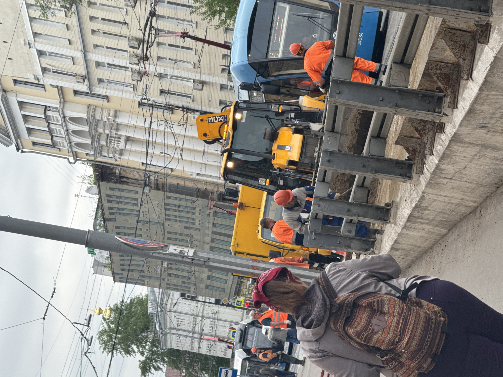 The image captures a street scene in Moscow, Russia, where construction or roadwork is underway. Workers in orange safety vests and hard hats are positioned around a yellow backhoe and a blue public transportation vehicle (possibly a bus or trolleybus with the number "31034" visible). They appear to be involved in some kind of maintenance or repair work, likely associated with the bridge infrastructure visible in the foreground.
A woman with a backpack and a red bandana on her head is visible closer to the camera, suggesting that pedestrians are sharing space with the construction activity. In the background, buildings with classic Russian architecture can be observed, as well as a church with a golden dome, alluding to the city's historical and cultural context. Overhead wires suggest the presence of a trolleybus or tram line.
