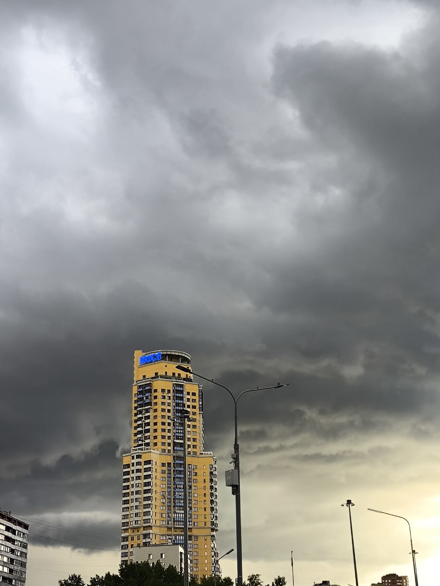 The image shows a tall, yellow building with blue lettering on top, likely an apartment building, set against a backdrop of heavy, dark storm clouds. The building has multiple floors and many windows. In the foreground, there are several street lights and some greenery, suggesting a street or urban area. Other buildings are visible in the distance. The location appears to be in an urban environment, specifically a city.