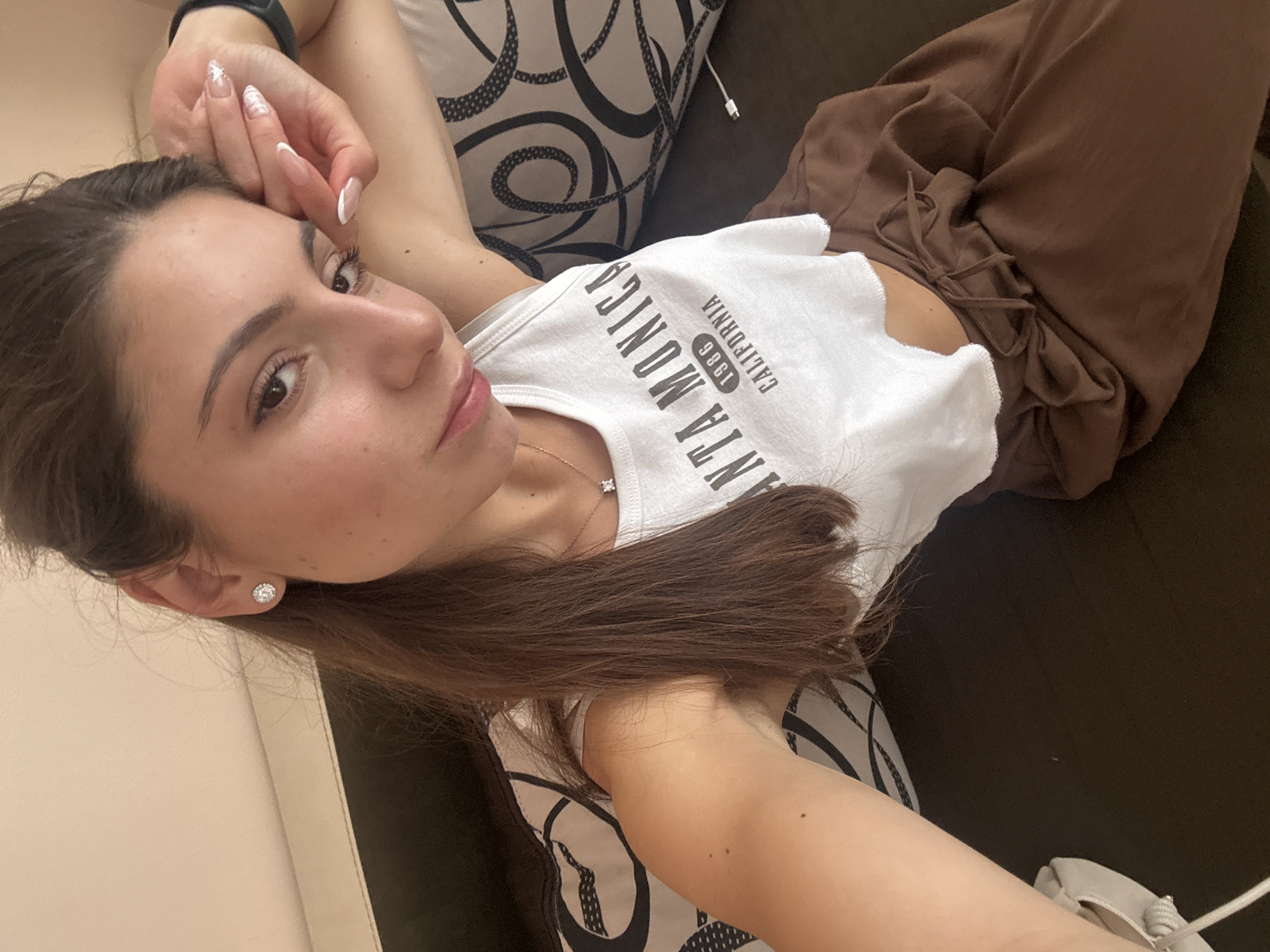 A young woman with long, dark brown hair, wearing a white crop top with "SANTA MONICA 1986 CALIFORNIA" printed on it, and brown pants, is lying down on a dark brown sofa with patterned pillows. She is looking at the camera, with her left arm resting behind her head and her right hand near her face. She is wearing a silver necklace with a diamond pendant and diamond stud earrings. Her nails are painted with a French manicure. The scene appears to be indoors, possibly in a living room or bedroom. The lighting is soft and natural. The environment is a residential setting. There is no clear indication of the city of Varna, Bulgaria.
