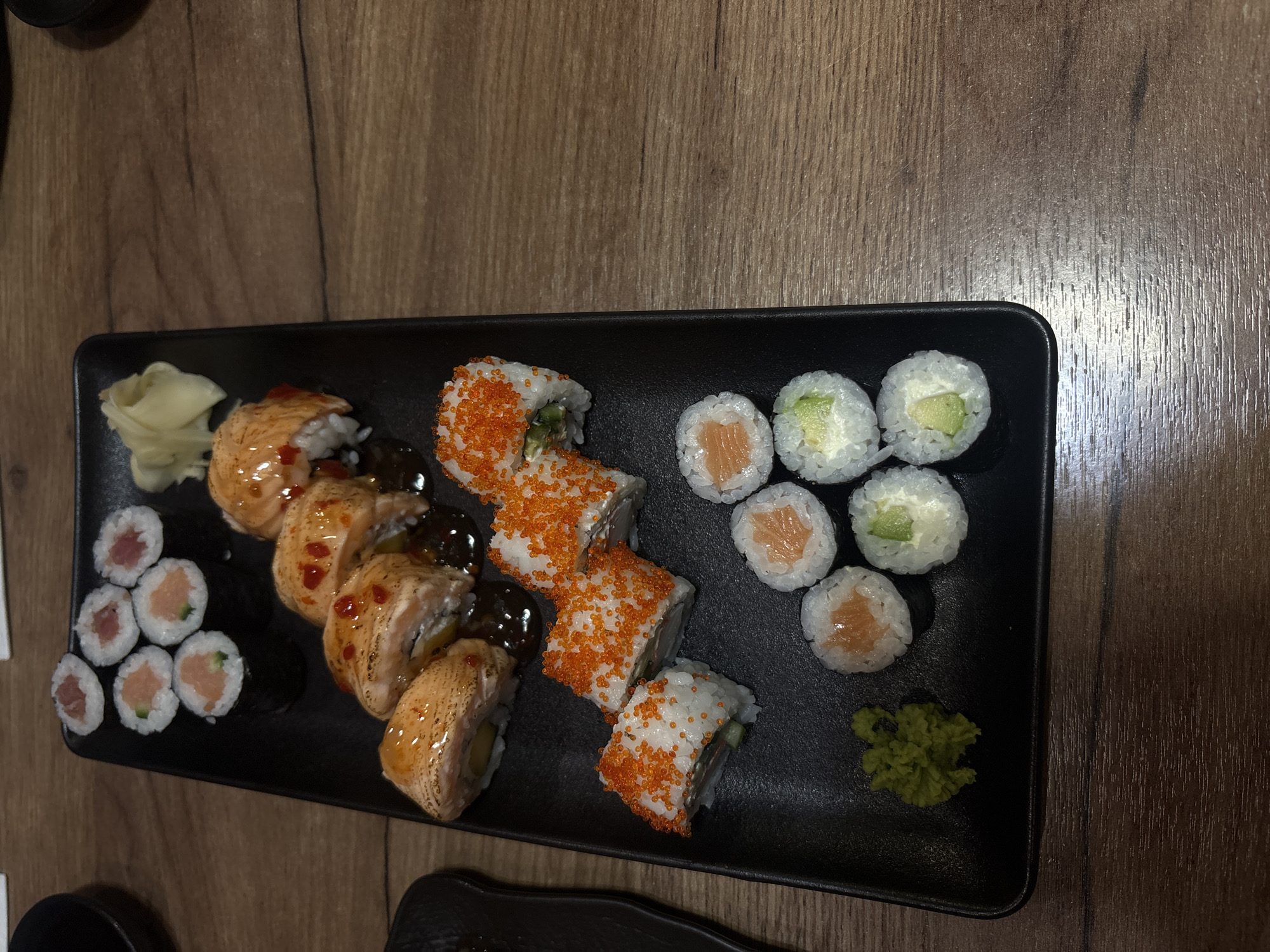 A top-down, medium shot shows a black rectangular plate of sushi on a brown wooden surface. The sushi includes tuna rolls, torched salmon rolls with a drizzle of dark sauce, California rolls with orange roe, salmon rolls, and avocado rolls with cream cheese. A dollop of ginger is on the left, next to the tuna rolls, and a blob of wasabi is on the right, next to the avocado rolls. The precise location in Varna, Bulgaria, cannot be determined from the image. No people are visible.