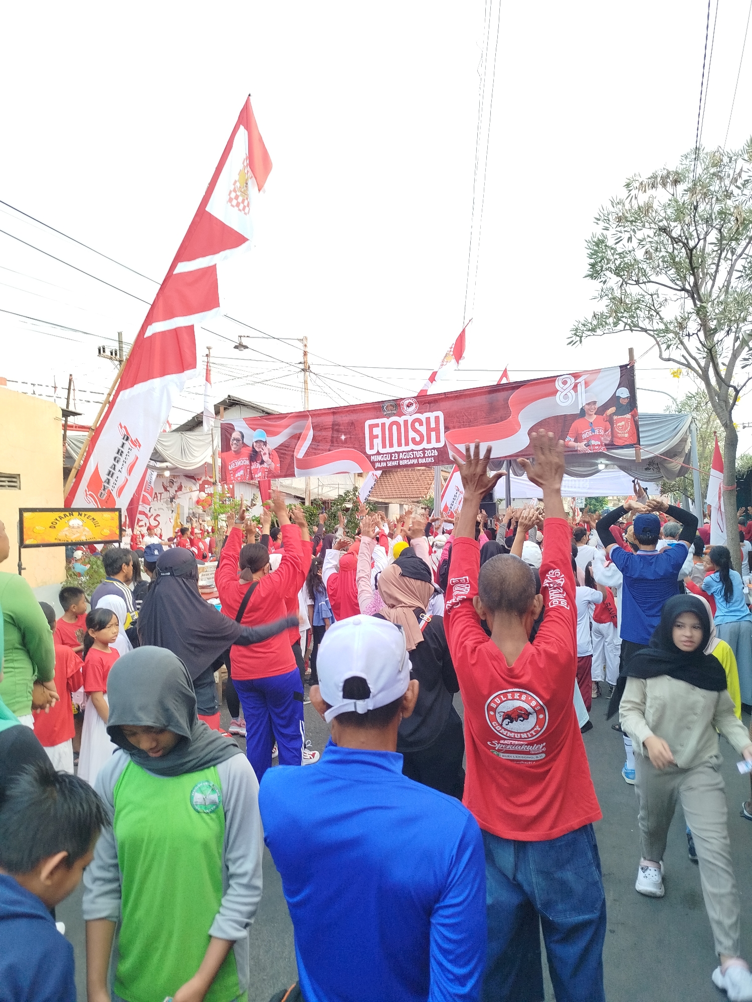 The image captures an outdoor event, likely a race or celebration, in Surabaya, Indonesia. In the foreground, a group of people are gathered, many wearing red shirts and some with head coverings. Their arms are raised in the air, suggesting excitement or participation. A banner prominently displays "FINISH" along with "MINGGU 23 AGUSTUS 2024" and "JALAN SEHAT BUKERS". The banner also features an "81" and images of two individuals.
Further back, more people are visible, and several Indonesian flags, red and white, are displayed. One large flag is positioned prominently on the left side of the frame. In the background, buildings and trees are present, indicating an urban environment. The overall scene suggests a festive and communal gathering.