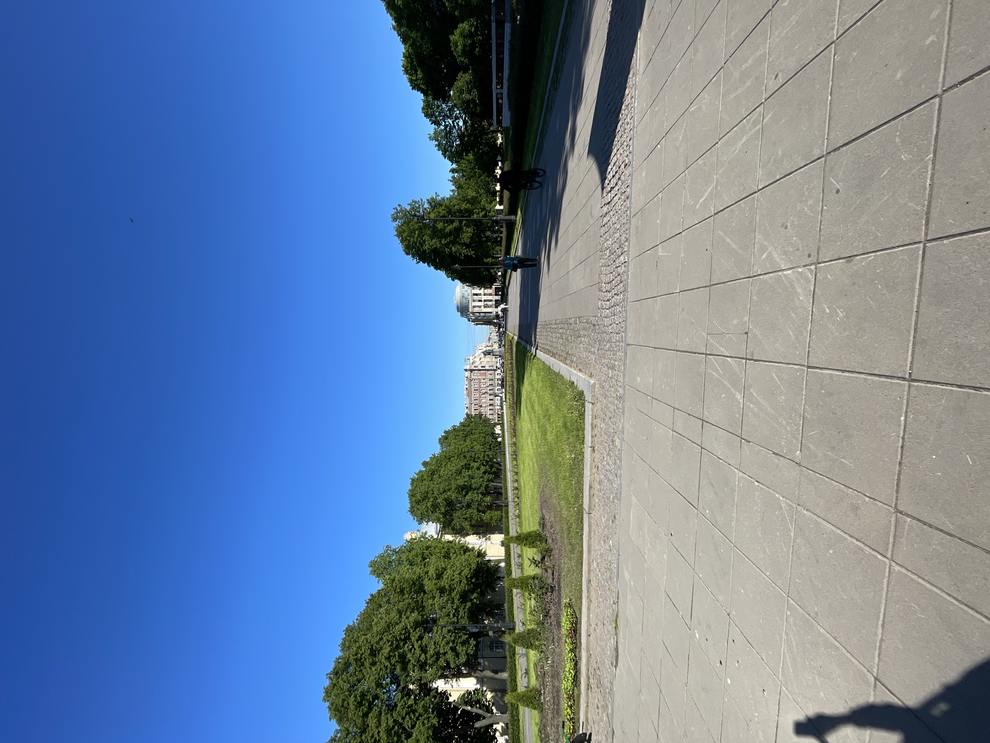 The image is oriented sideways, rotated 90 degrees clockwise. It depicts a sunlit urban park or wide boulevard in Saint Petersburg, Russia, under a clear, bright blue sky.
A broad paved walkway, composed of large square grey tiles, dominates the right side of the frame, extending from top to bottom. This path is bordered by a strip of darker cobblestones. The tiles show some scuff marks and dirt. The shadow of the photographer, holding a device, is cast onto the bottom-most right corner of this path.
To the left of the path, a vibrant green lawn stretches out, running parallel to the walkway. Within the grassy area, rows of smaller, neatly trimmed bushes are visible. Further to the left, a dense line of mature, leafy green trees creates a lush boundary, with some streetlights partially visible among them. In the distance, beyond the trees, multi-story buildings with classical architectural elements, including a prominent dome, rise against the horizon. Some buildings appear lighter, others are reddish-brown.
Two individuals are discernible in the scene: one person in dark clothing walks on the paved path further up the frame, while another, wearing lighter attire, is visible further down near the trees. A small, dark silhouette, possibly a bird or drone, is high in the clear blue sky in the upper left corner. A green electric scooter is partially visible on the grass near the edge of the paved path.
The strong sunshine casts long shadows from the trees and objects, indicating it is either morning or late afternoon on a clear, pleasant day. There is no visible text in the image.