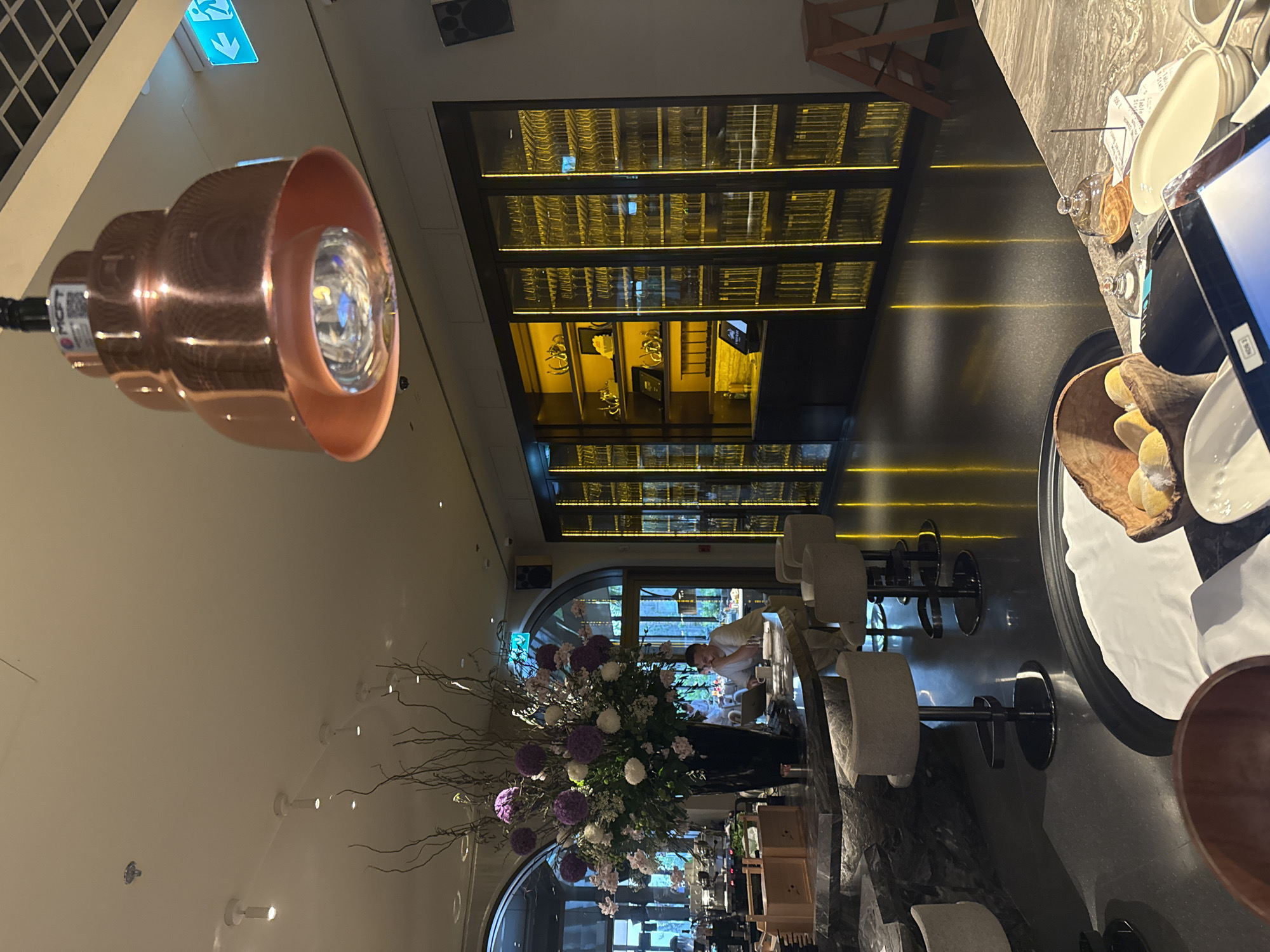 The image depicts the interior of a restaurant bar area in Dubai, United Arab Emirates. In the foreground, a marble countertop is visible with various items, including a wooden bowl containing yellow objects, small glass cloches, and a tablet device. Behind the counter, a man is seated at the bar, looking at a laptop with a cup on the counter. A large floral arrangement with purple and white flowers in a dark vase is prominently displayed on the left side of the bar. In the background, a long, illuminated display cabinet filled with glassware stretches across the wall. Bar stools are arranged around the counter. The lighting is warm and ambient, with a decorative copper pendant light hanging from the ceiling. An illuminated exit sign with an arrow pointing left is visible on the upper right wall.