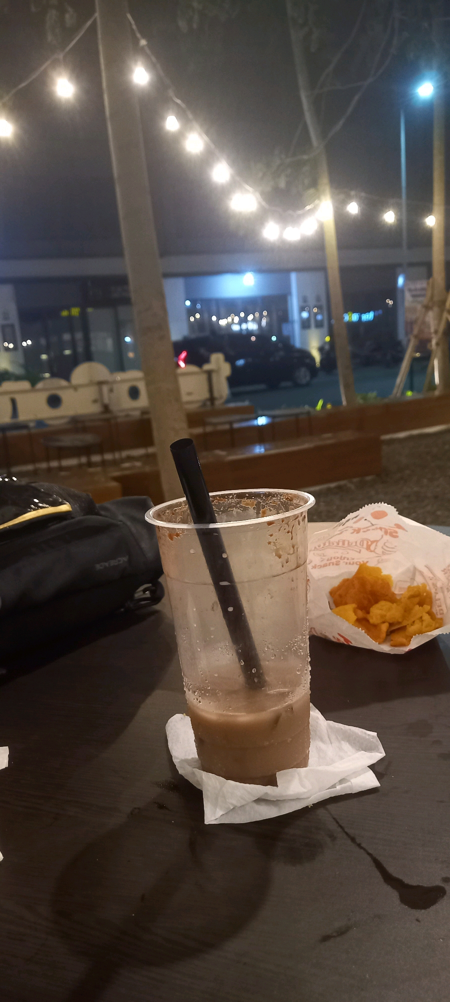 The image captures an outdoor evening scene at a commercial establishment in Sidojangkung, Indonesia. In the foreground, a partially consumed beverage in a clear plastic cup with a black straw rests on a dark, wooden table, accompanied by a white napkin. To the right of the cup, a paper bag filled with fried food items, possibly crackers or chips, is visible. A black bag is positioned to the left of the cup. The background reveals string lights illuminating trees and a building with other patrons and vehicles. The lighting suggests a relaxed, social atmosphere.