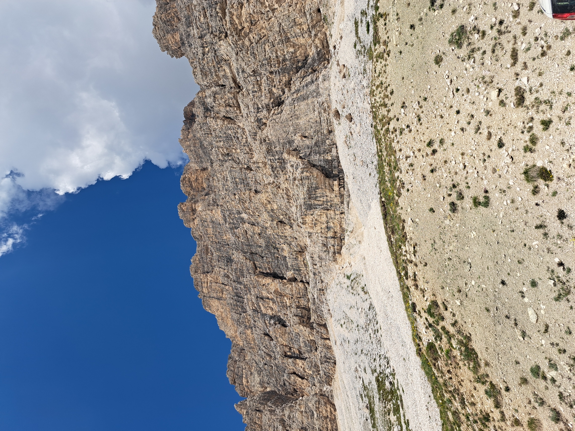 The image captures a dramatic, vertical expanse of a rocky mountain face, likely a cliff. The rock is layered and textured, with deep fissures and crags that create shadows, suggesting the sun is positioned to highlight these features. To the left, a vibrant blue sky dominates, punctuated by a few scattered white clouds. The ground at the base of the cliff appears to be a rocky, arid terrain with sparse vegetation, including small bushes and tufts of grass.
The perspective is looking upwards at the mountain, emphasizing its sheer scale and ruggedness. The orientation of the photo is vertical, which further accentuates the height of the cliff. The lighting suggests it is daytime, likely late morning or afternoon, with clear weather conditions except for the clouds. There are no people or discernible objects on the cliff face itself, but a small portion of a white vehicle with a red accent is visible at the very top right edge of the frame, suggesting human presence or access to the area. The overall impression is one of a vast, natural landscape, highlighting the raw power and beauty of the mountains.