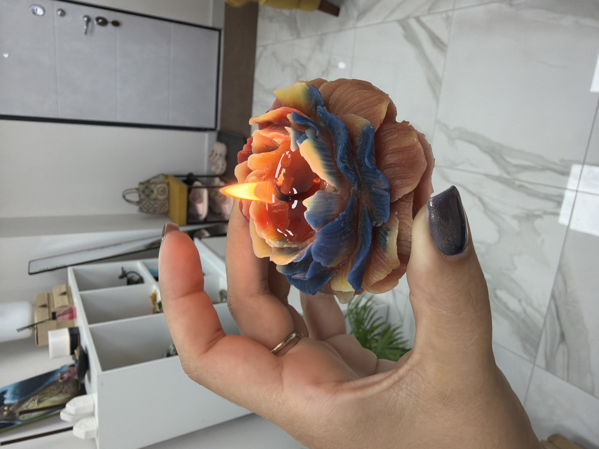 A person's hand holds a lit, multi-colored rose-shaped candle. The candle has petals in shades of peach, blue, and yellow, with a molten wax center where the flame burns. The hand is positioned in the foreground, with the thumb and index finger supporting the candle. The background shows an indoor environment with a white shelving unit, a door, and a yellow chair. The floor is tiled with a marble pattern. There is no information to suggest this scene is located in Petropavlovsk-Kamchatsky, Russia.